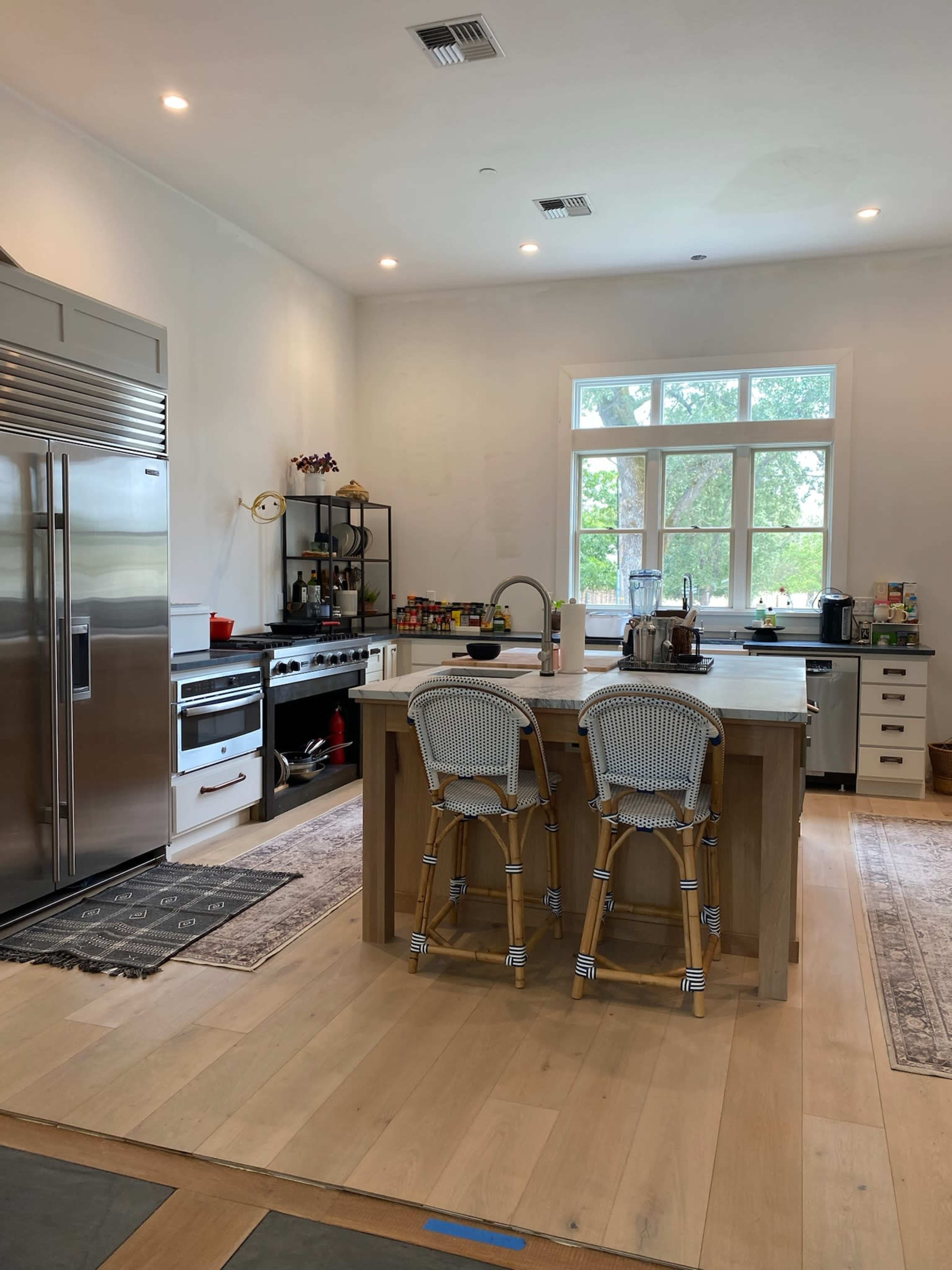 A modern kitchen features stainless steel appliances, a central island with two bar stools, and a large window overlooking greenery.