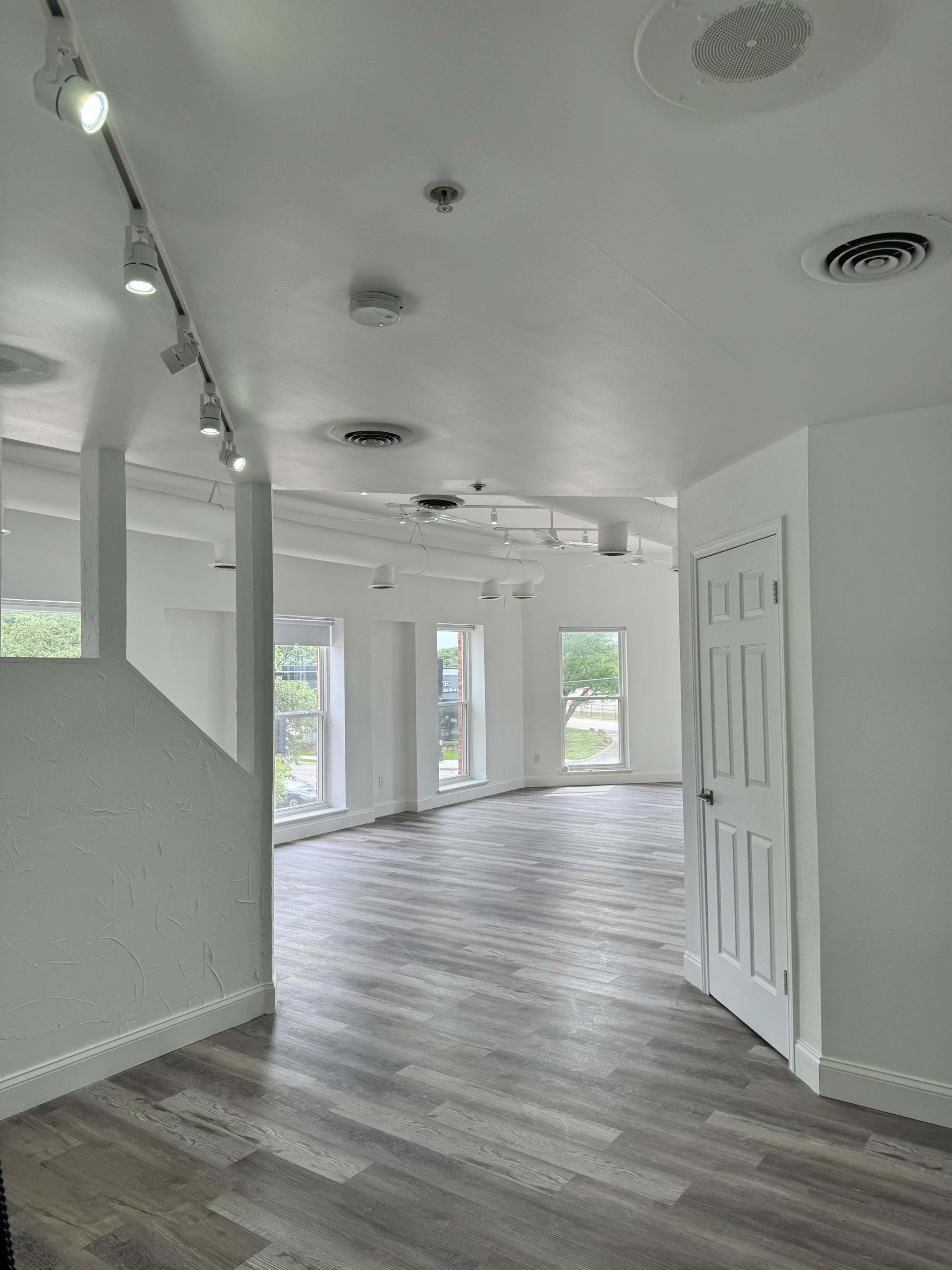The image shows a spacious, empty interior with light-colored walls and wooden flooring, featuring large windows and track lighting.