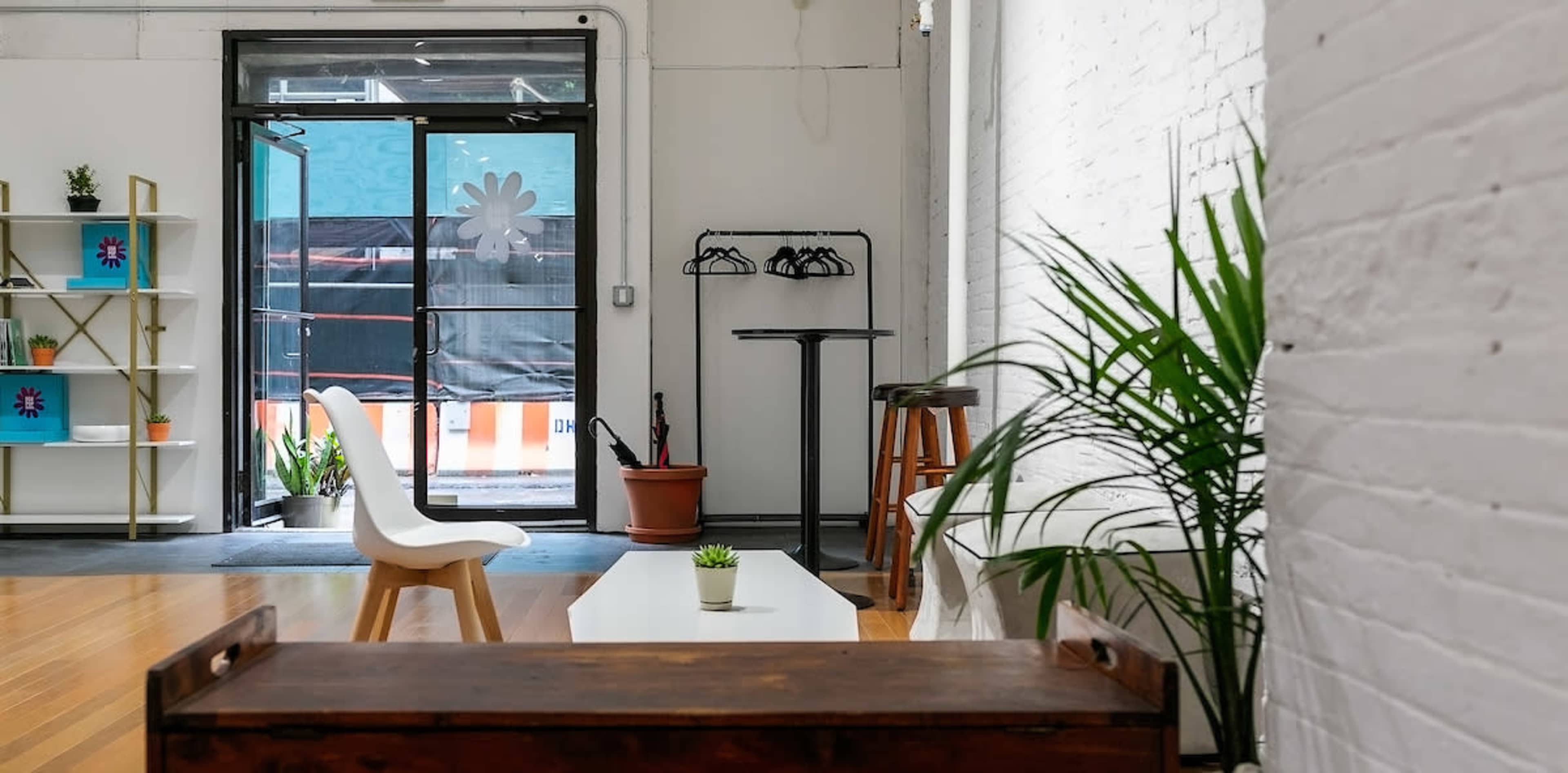 The scene shows a brightly lit, minimalist interior with a white brick wall, a small table, a chair, and a potted plant near a large door leading outside.