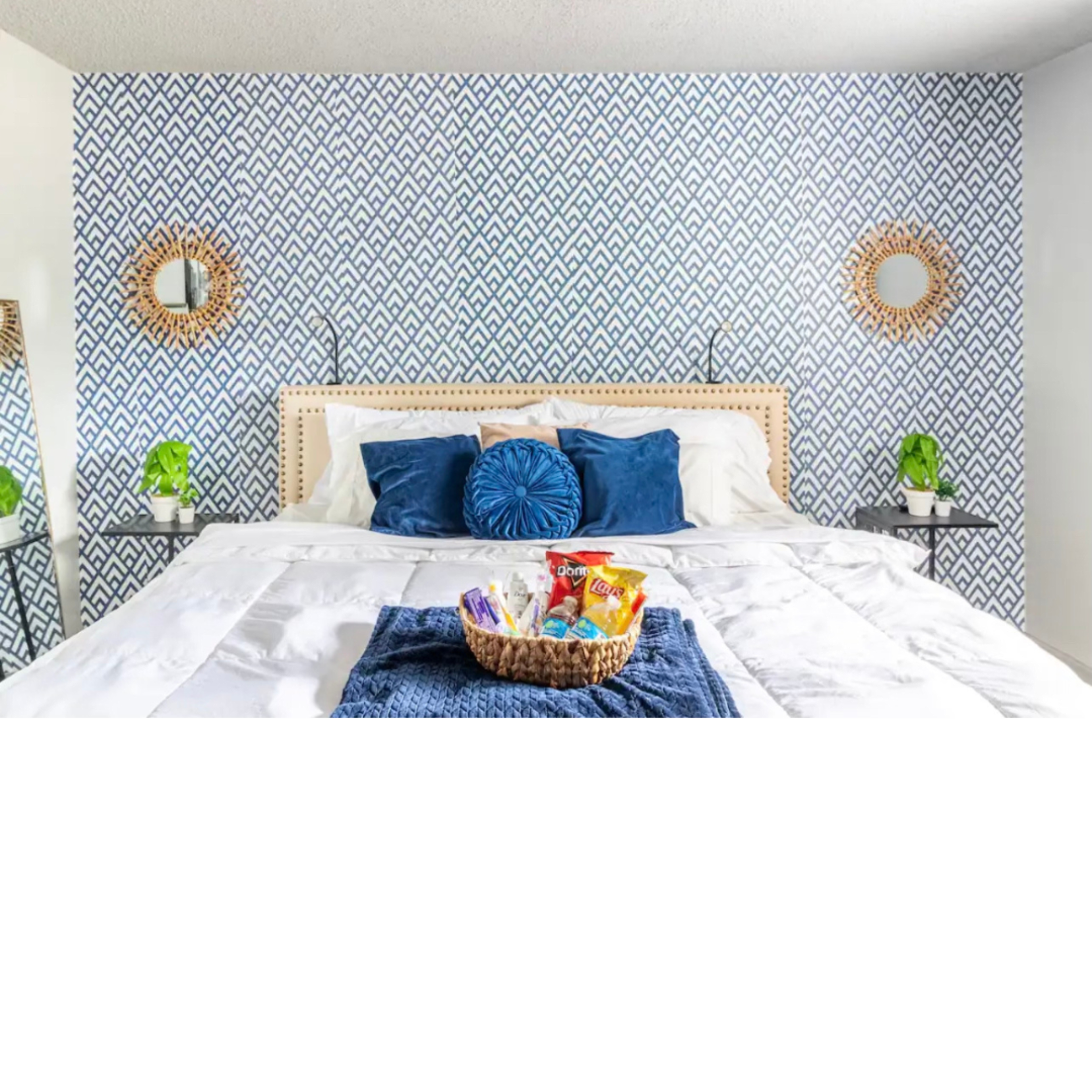 A neatly arranged bedroom features a large bed with white linens, blue decorative pillows, and a woven basket filled with snacks, set against a patterned blue and white wall.