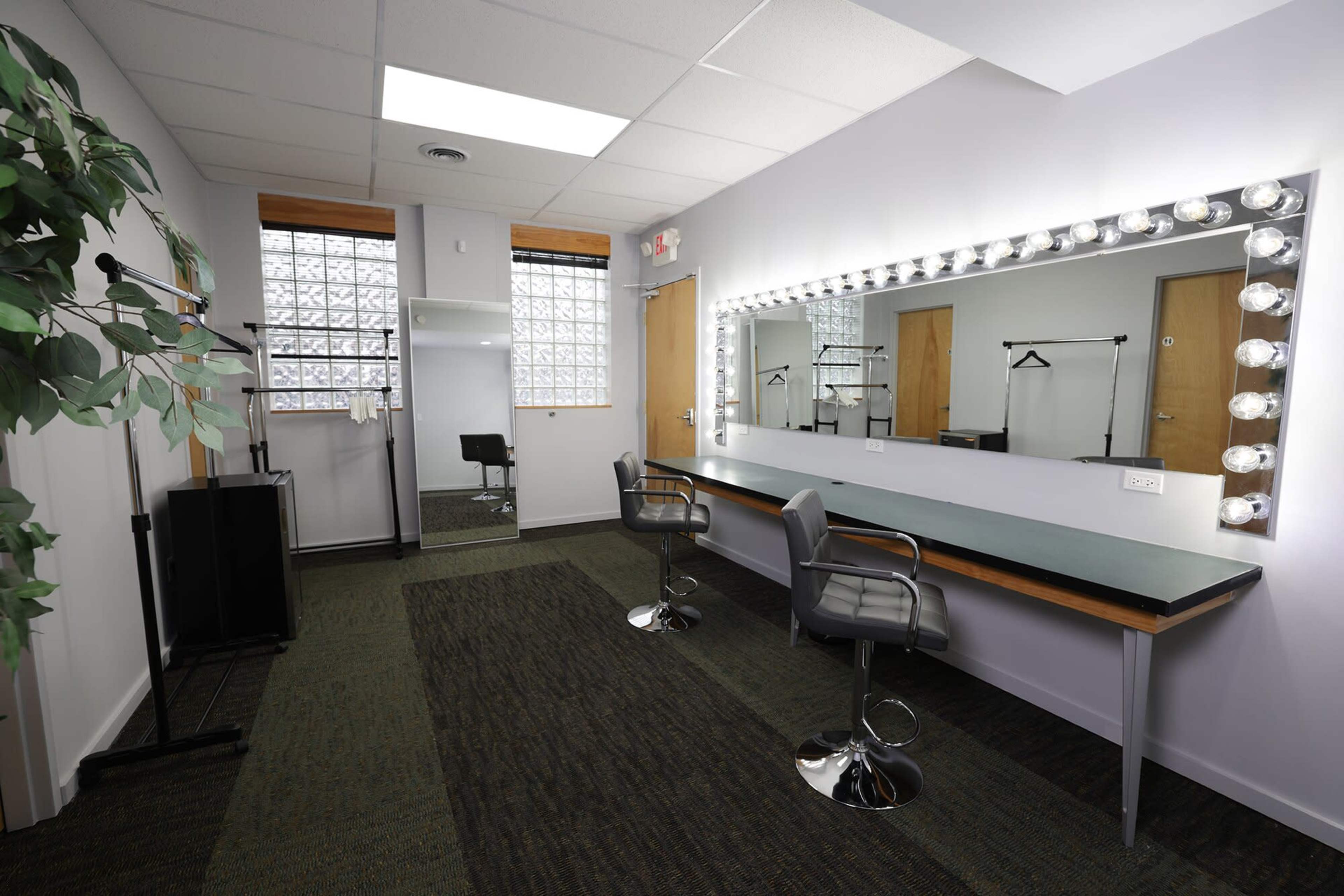 A dressing room with a large illuminated mirror, two barstools, and a green carpeted floor, along with a plant and clothing racks in the background.