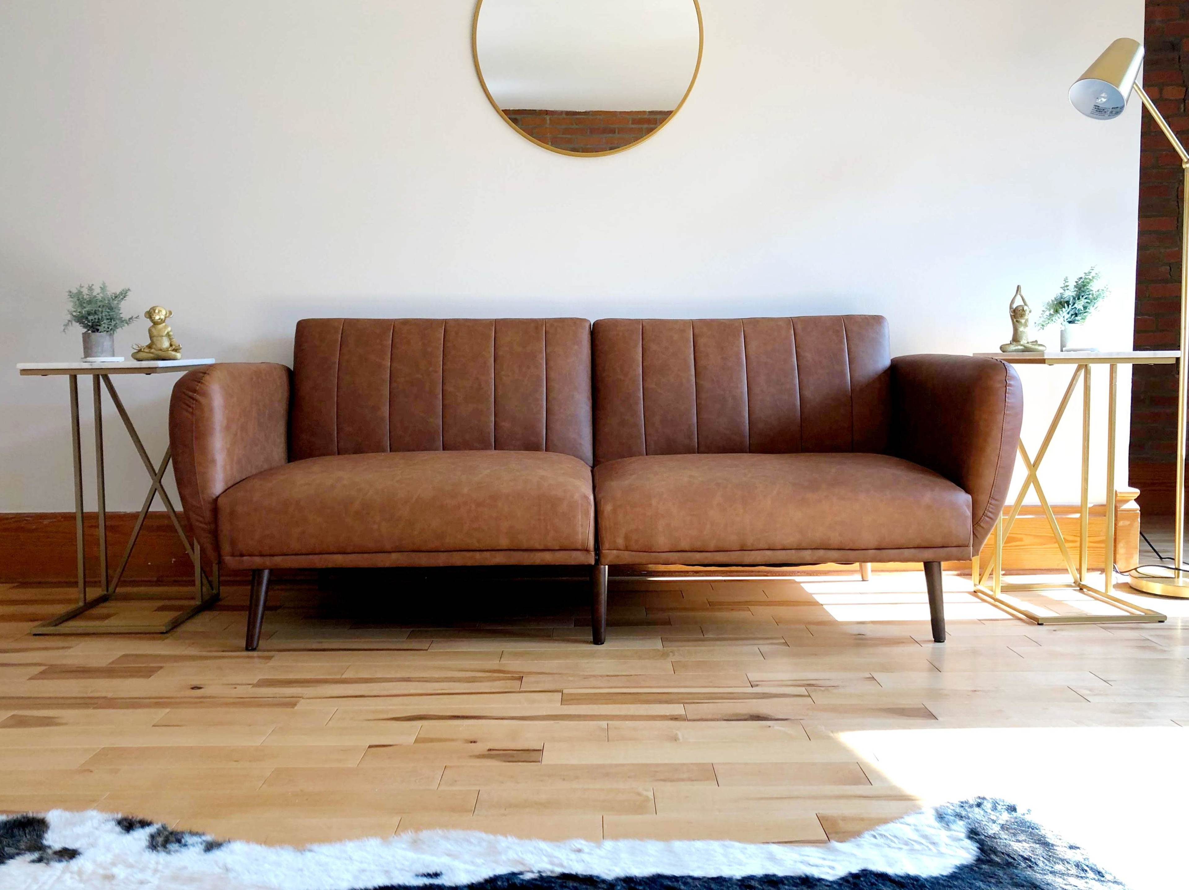 A brown leather sofa is positioned between two side tables with decorative items, in a room with a round mirror and wooden flooring.