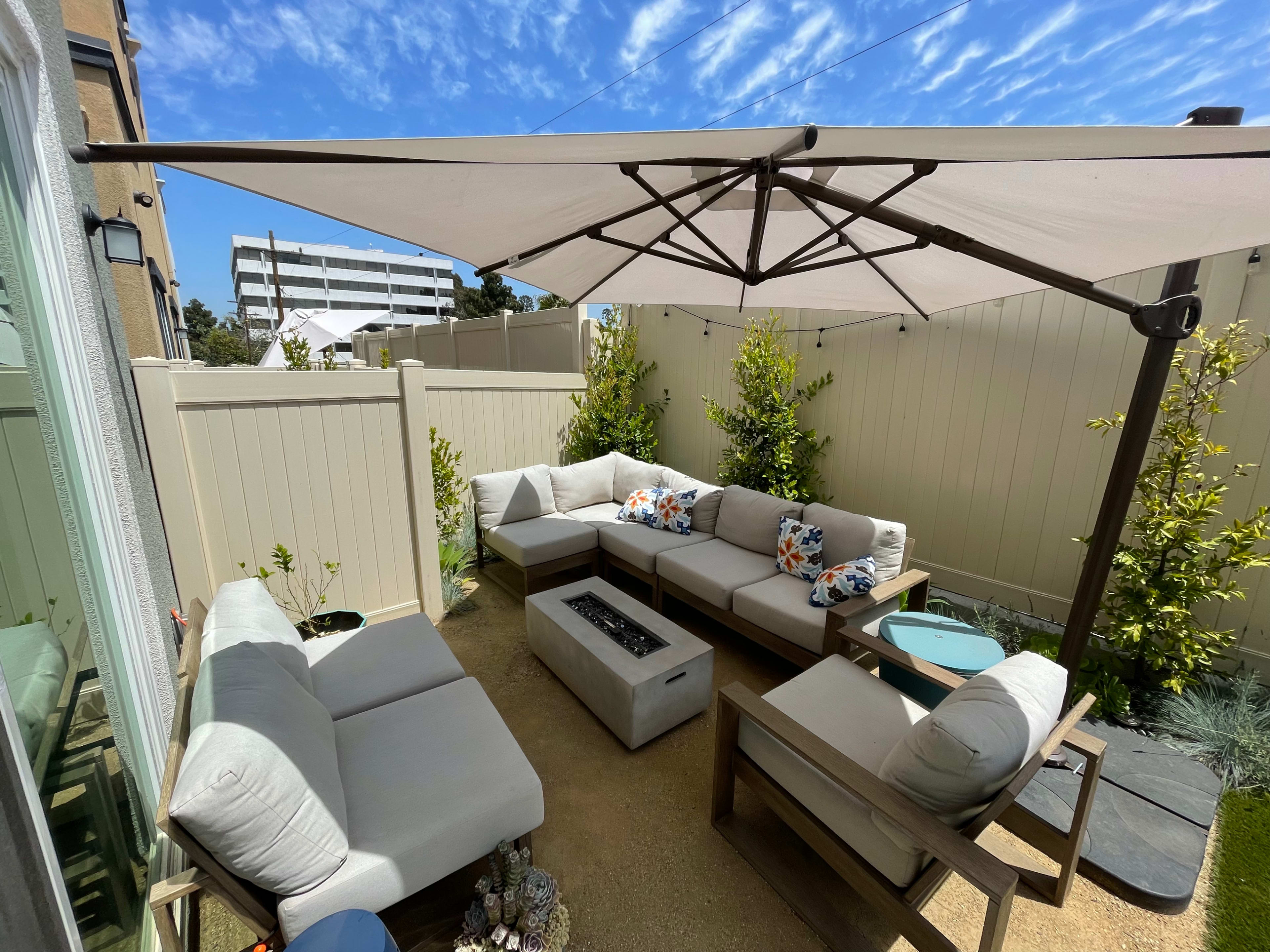 A patio area features a sectional seating arrangement under a large umbrella, with a central fire pit and potted plants surrounding it.