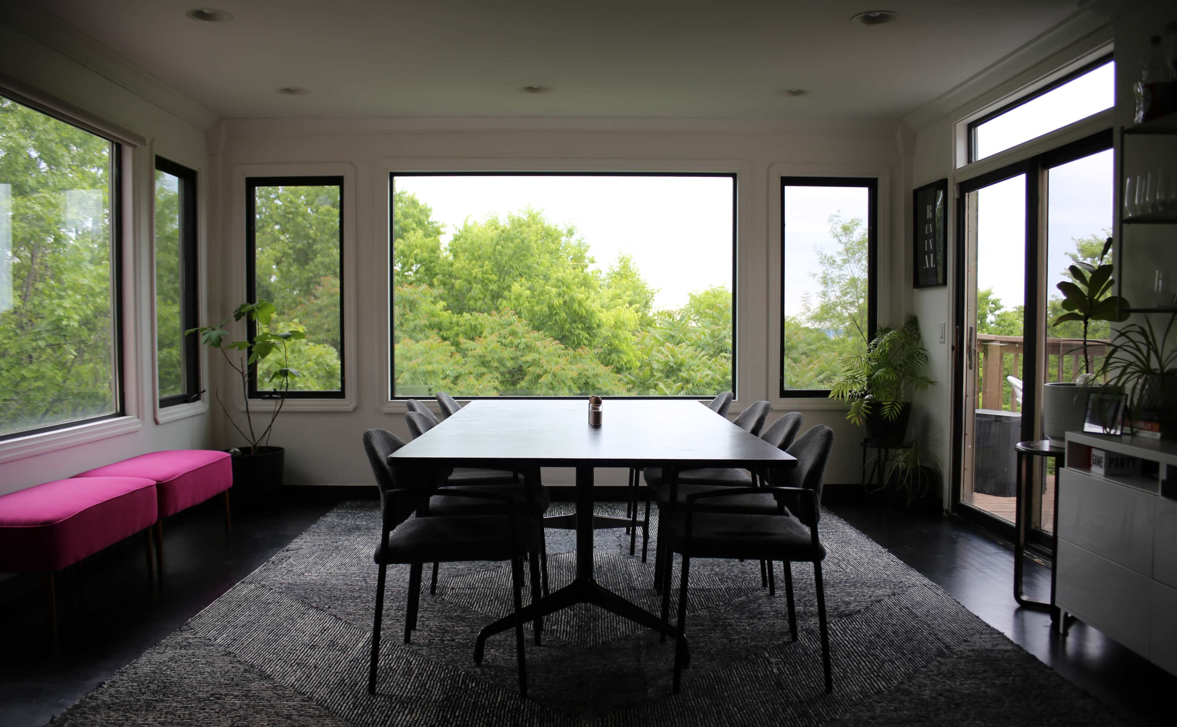 A modern dining room features a large table surrounded by chairs, with large windows showcasing greenery outside.