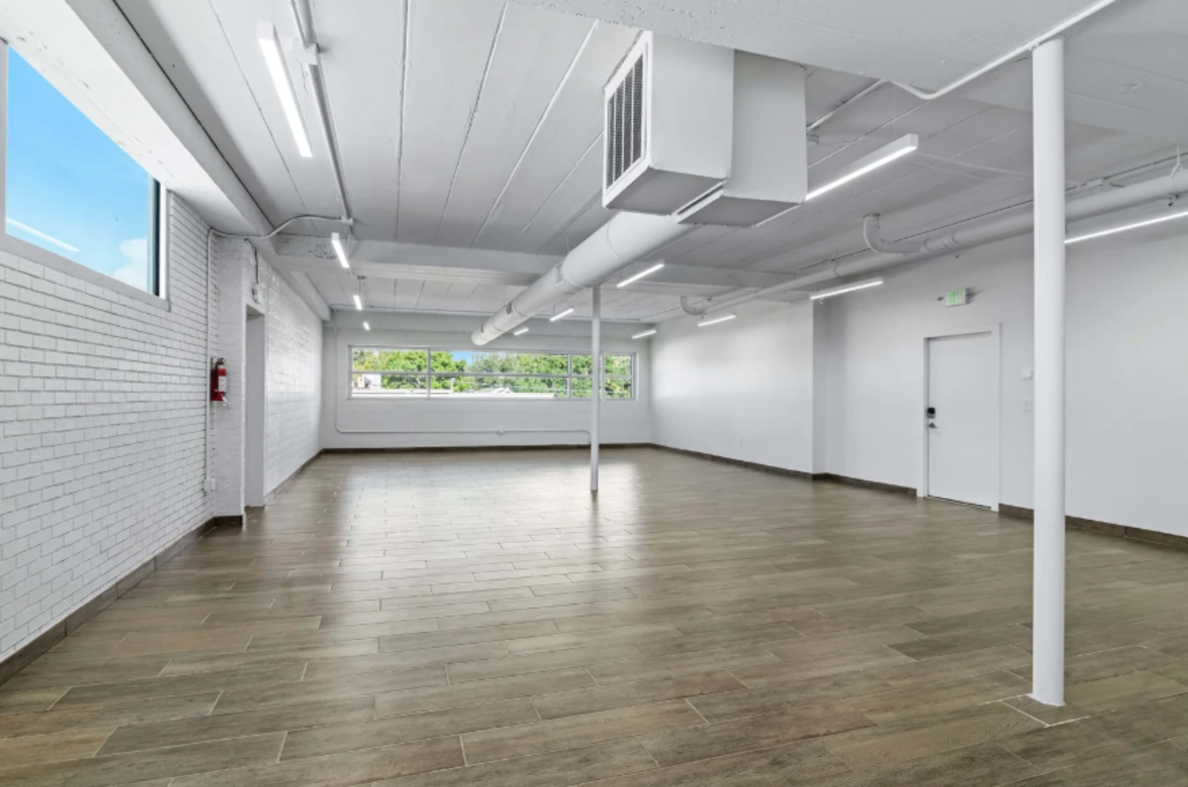 The image shows an empty, well-lit rectangular room with white walls and a long wooden floor.