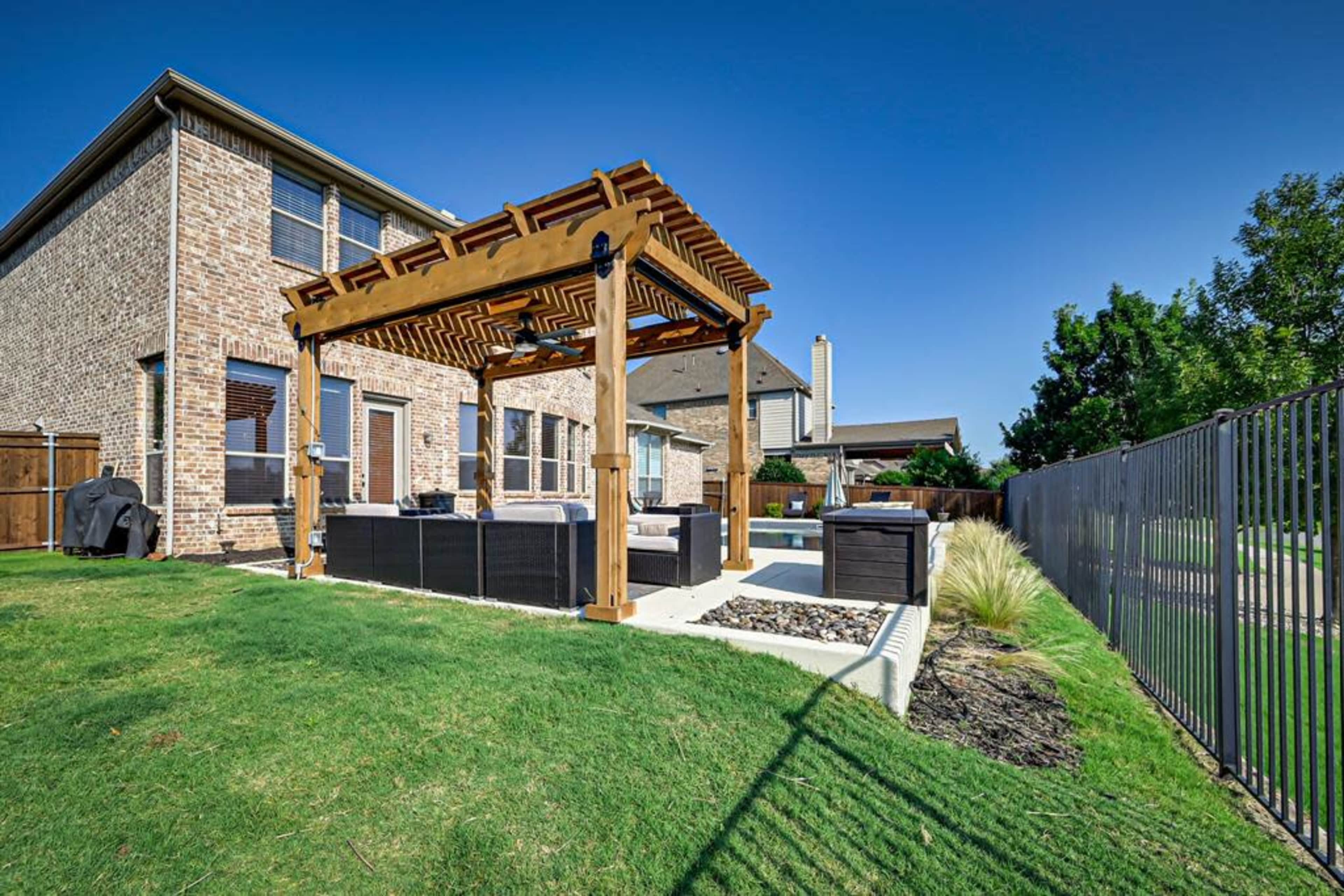 A wooden pergola provides shade over a seating area next to a hot tub in a grassy backyard.