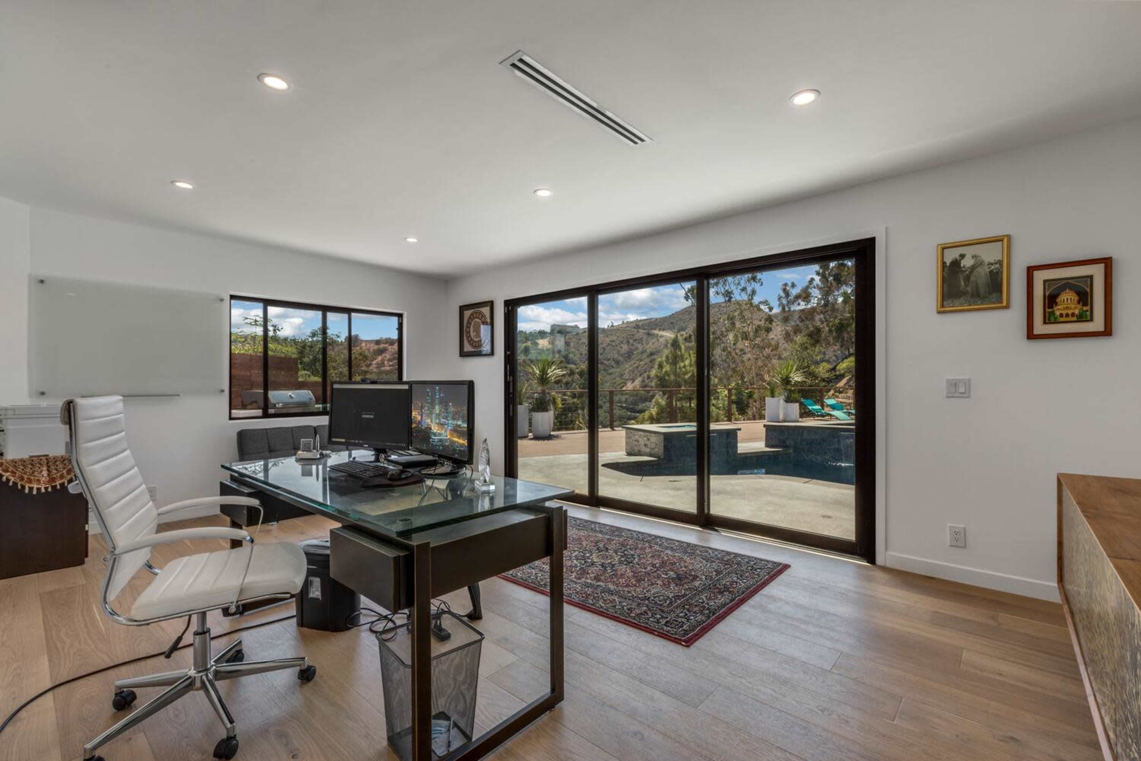 A modern office space features a glass desk with a computer, overlooking a patio and pool area through large sliding glass doors.