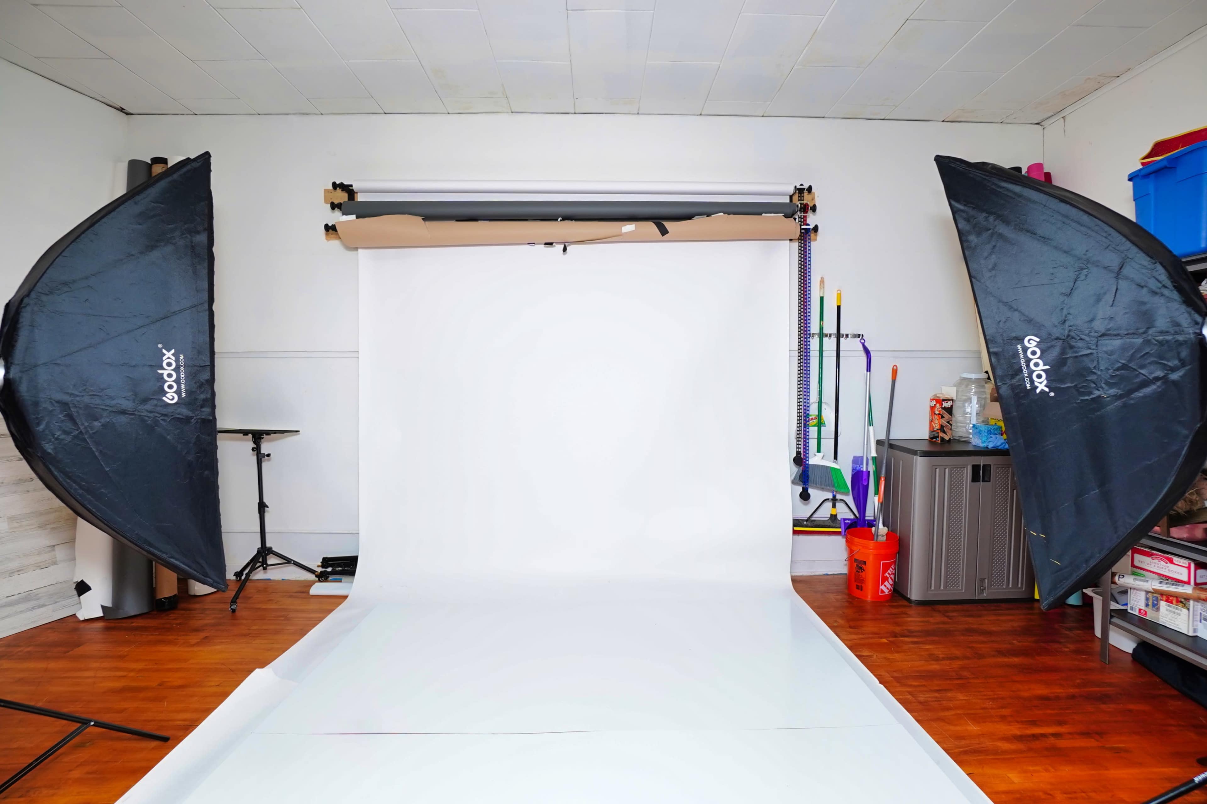 The image shows a photography studio setup with two softbox lights, a seamless backdrop, and various equipment in the background.