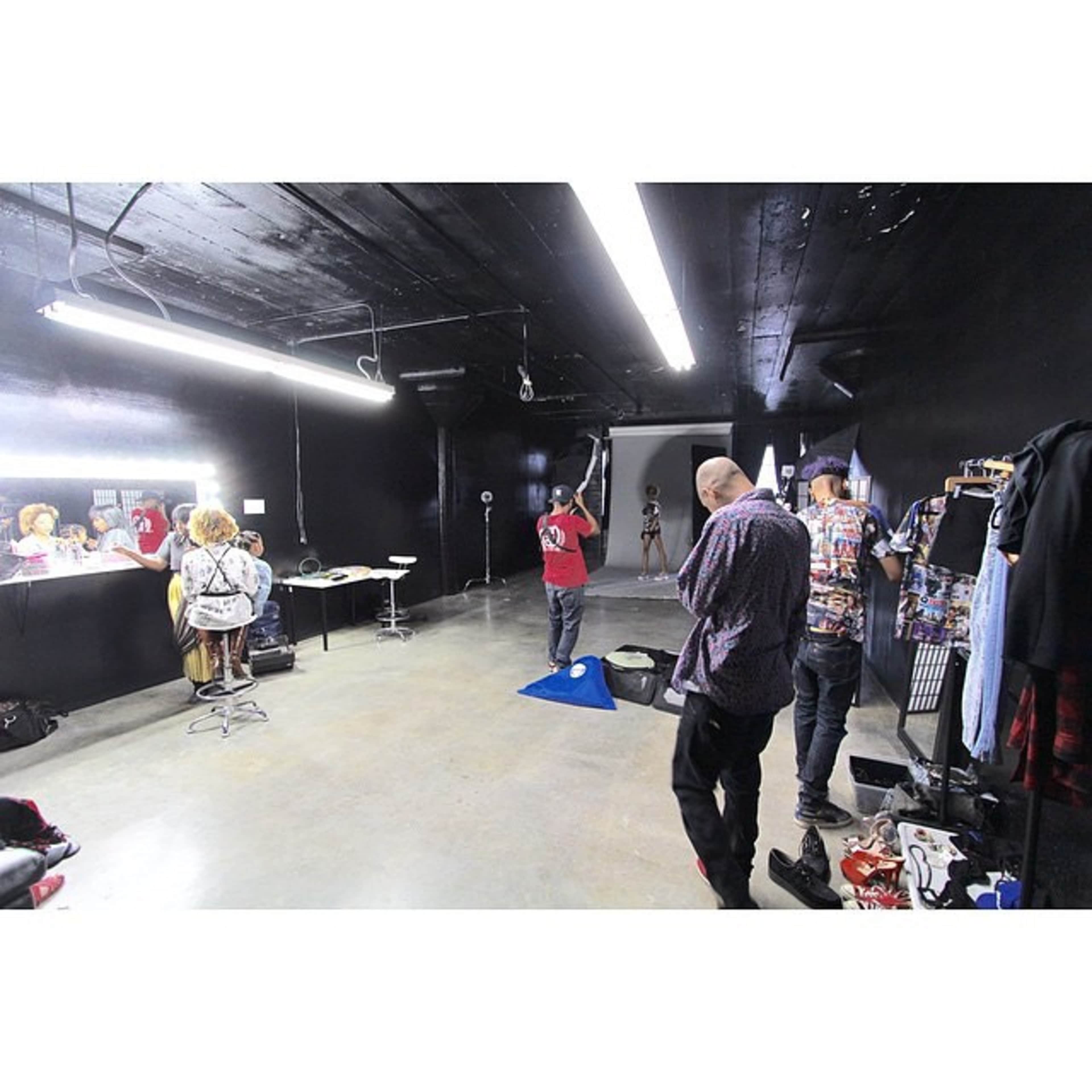 A photography studio with black walls featuring a model posing in front of a backdrop while others adjust lights and equipment.