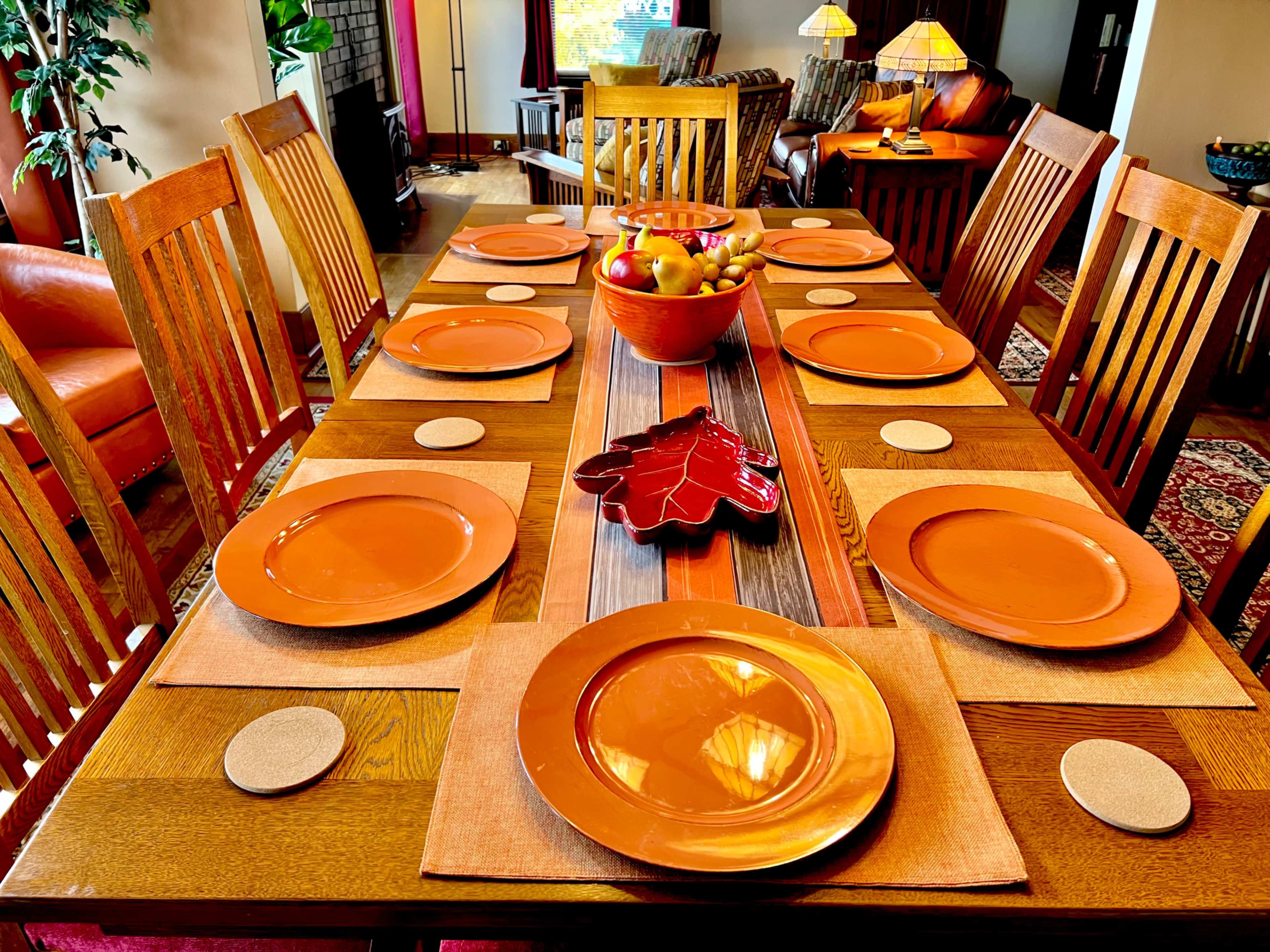 A wooden dining table is set with orange plates and placemats, featuring a centerpiece of fruit in a decorative bowl.