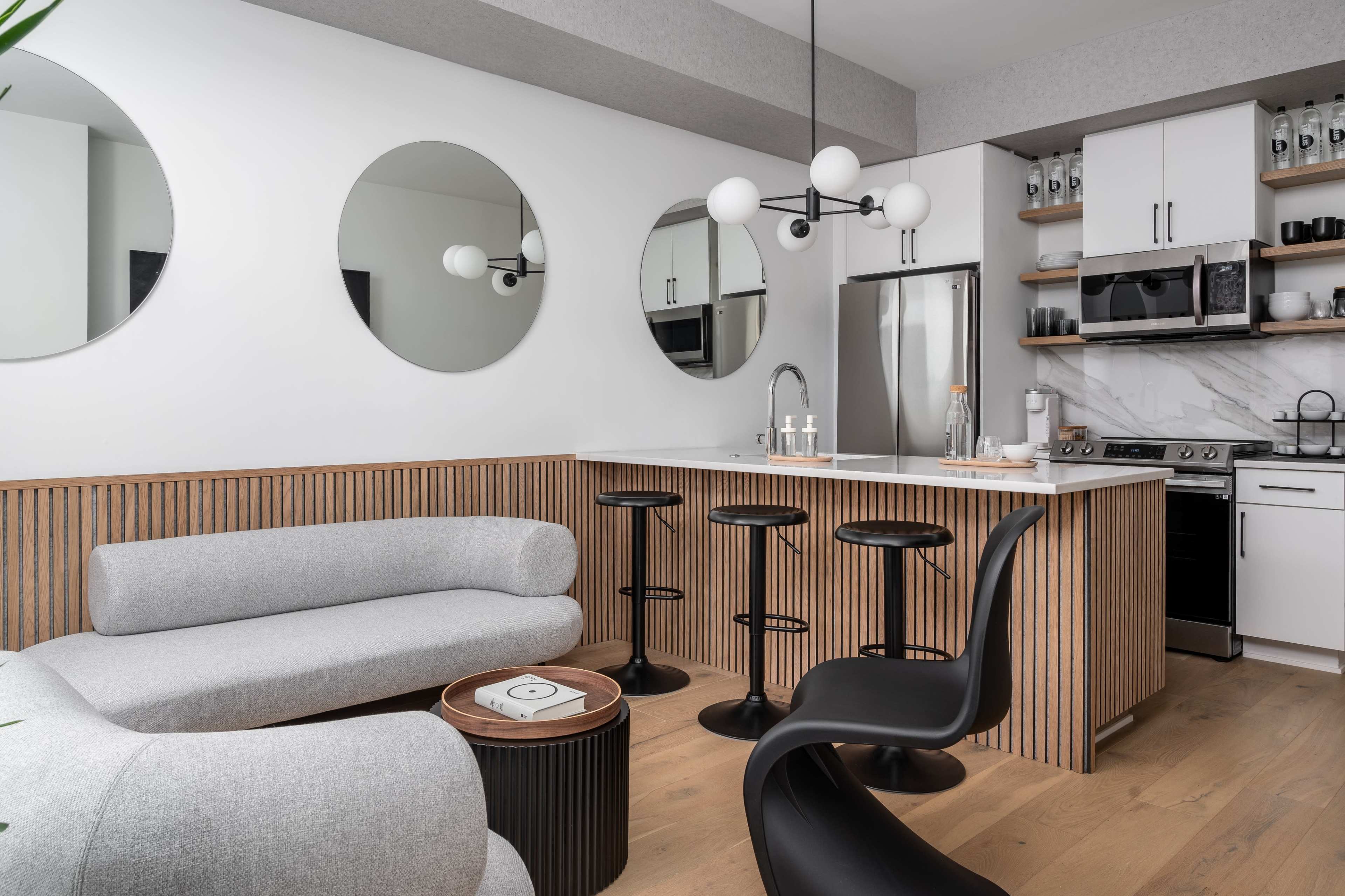 The image shows a modern kitchen and living space with a curved gray sofa, black seating at a kitchen island, and circular mirrors on the wall.