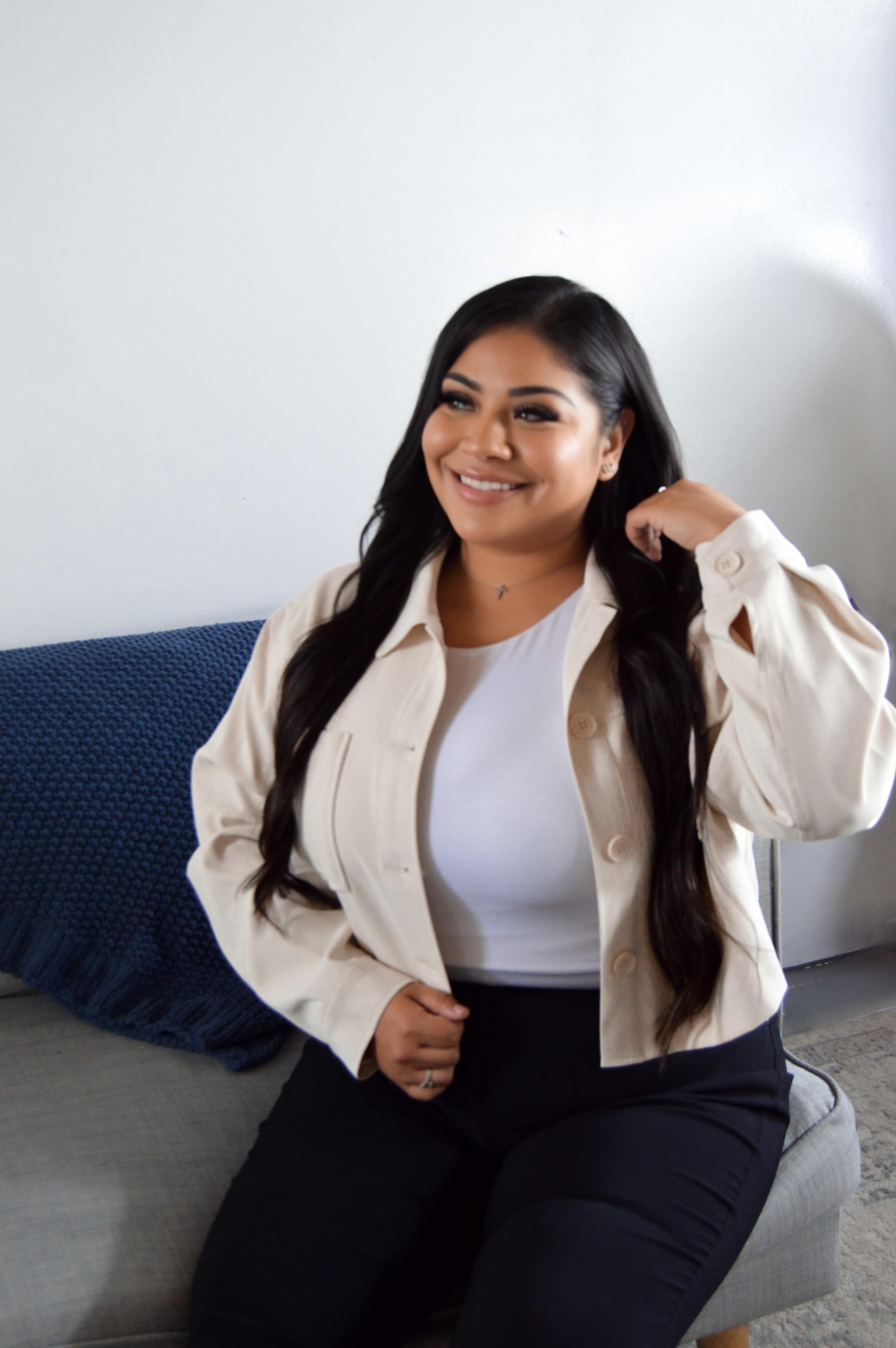 A woman with long dark hair is sitting on a couch and smiling in a well-lit room.