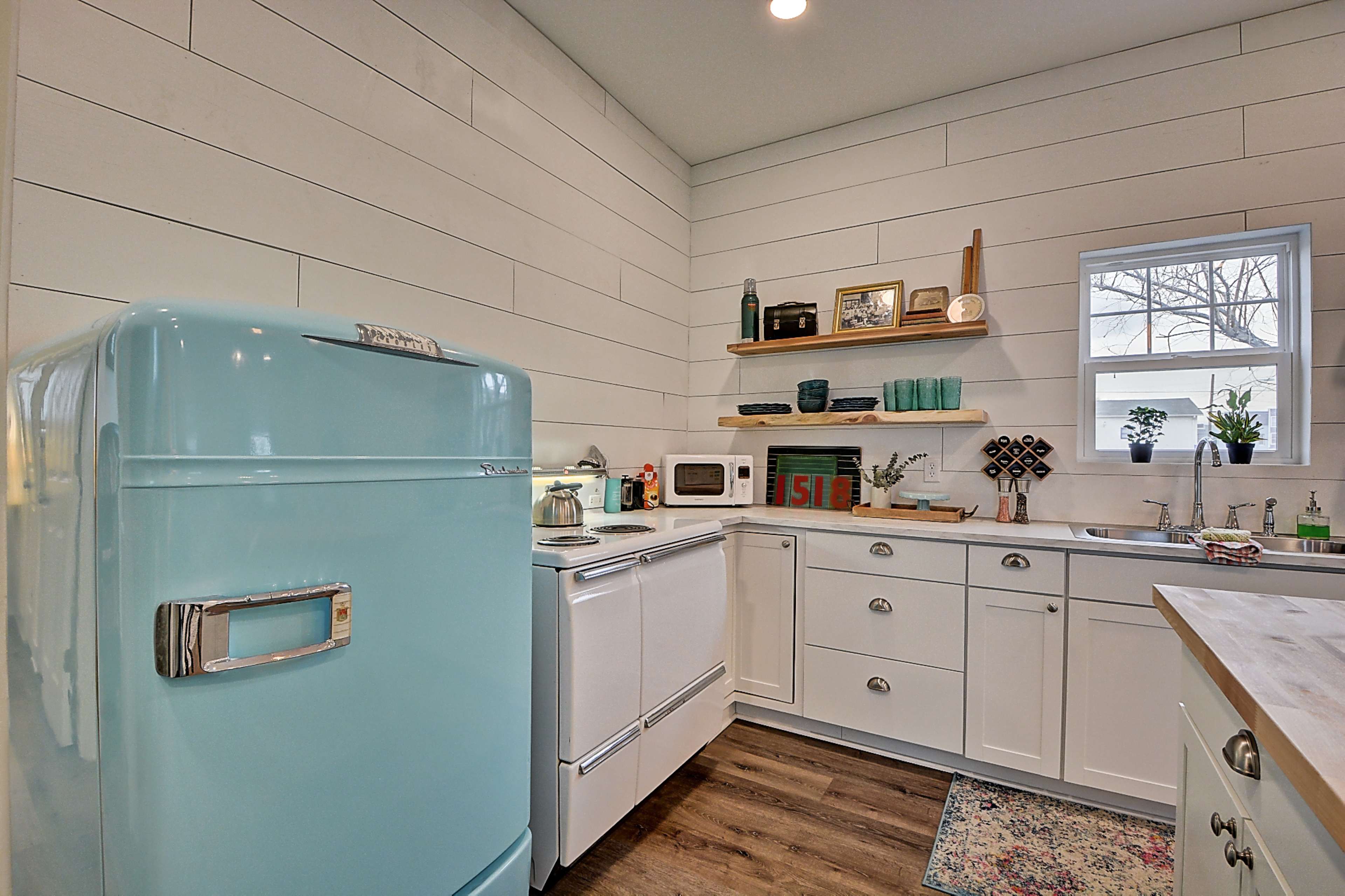 A bright kitchen features a retro turquoise refrigerator, white cabinets, and wood countertops, with a window providing natural light.