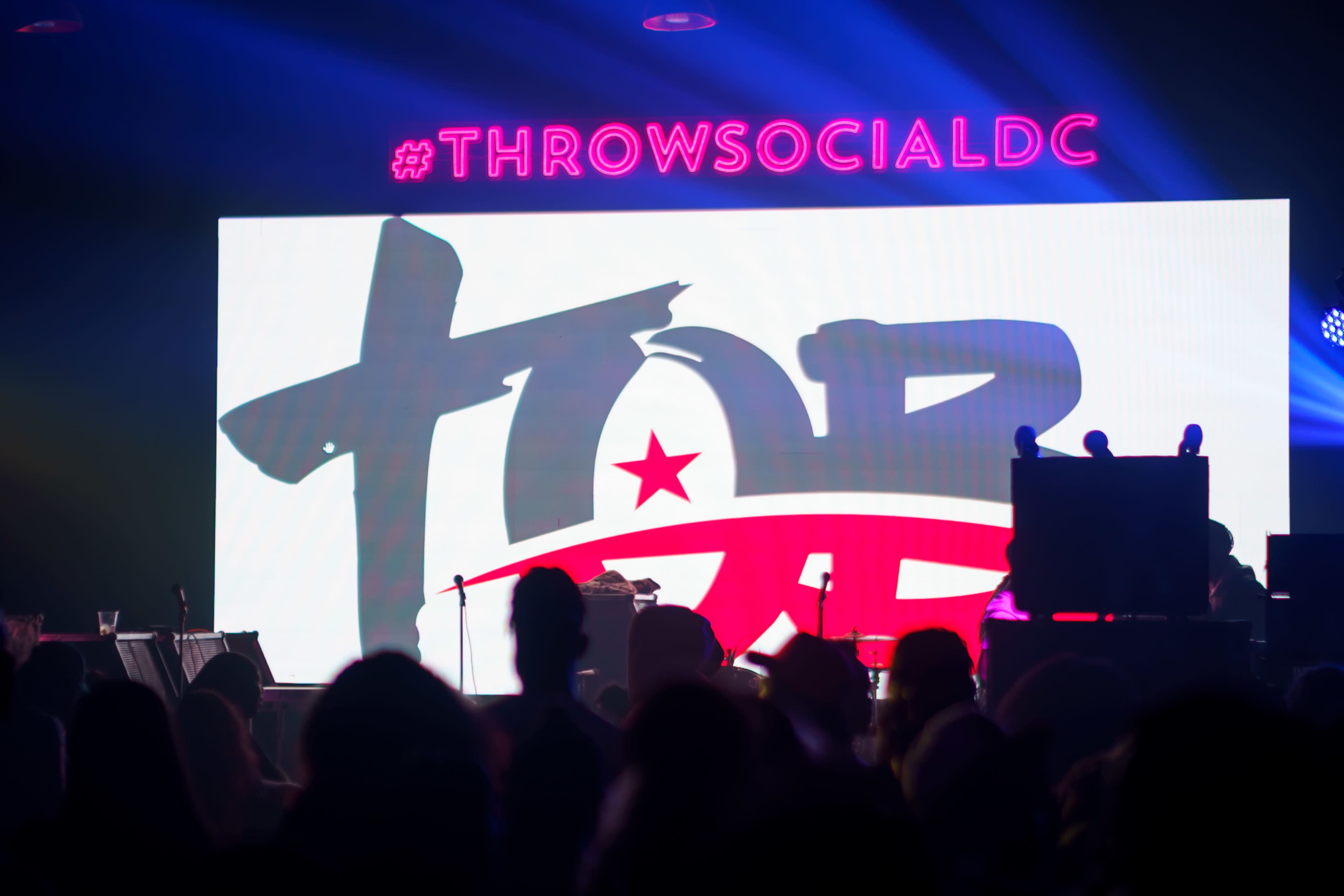 A large illuminated screen displays the hashtag #THROWSOCIALDC alongside a logo featuring the letters "TOP" with a star, while silhouettes of an audience can be seen in the foreground.