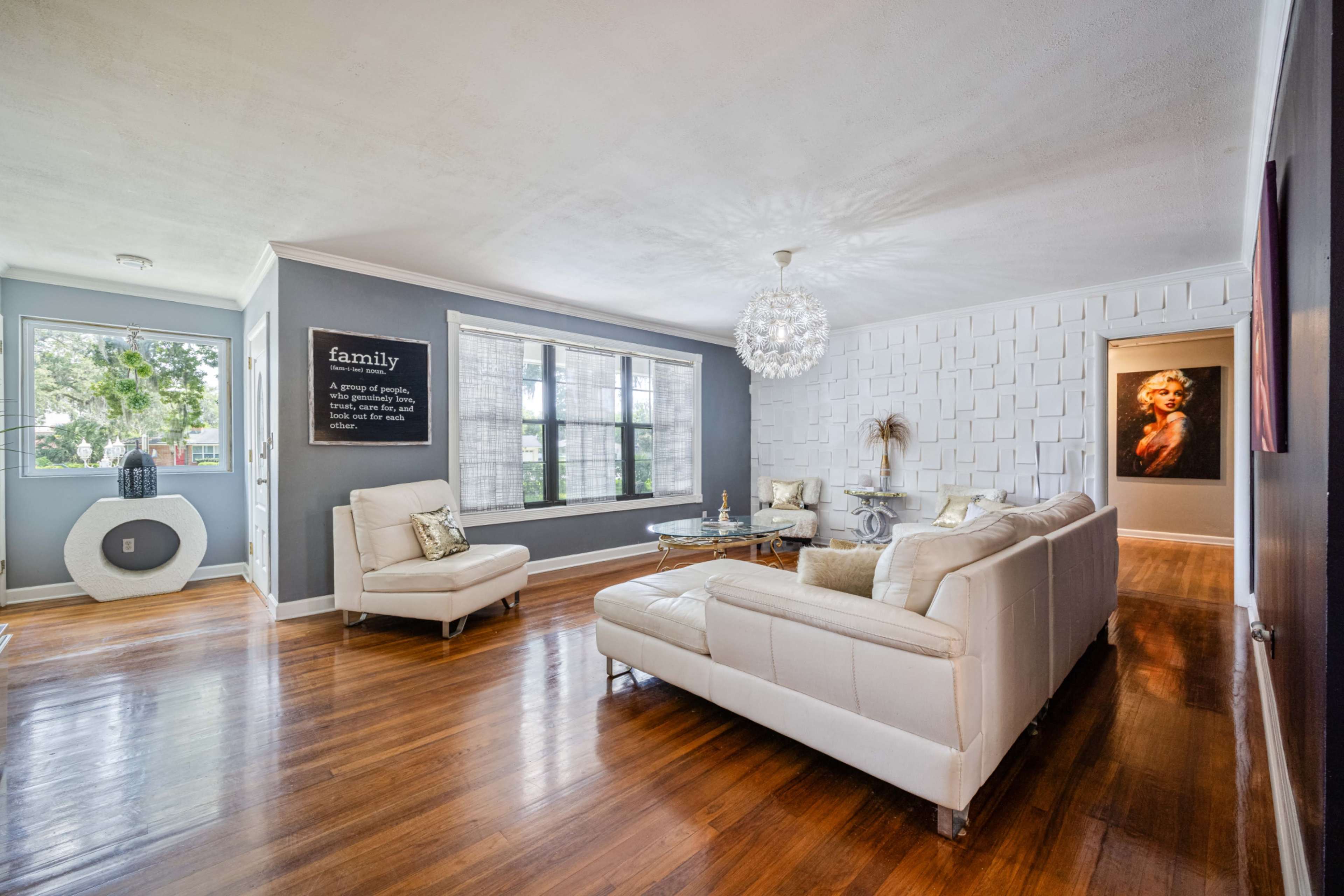 A modern living room features a white leather sofa, a glass coffee table, and a large window overlooking greenery, with a decorative wall art piece on the opposite side.