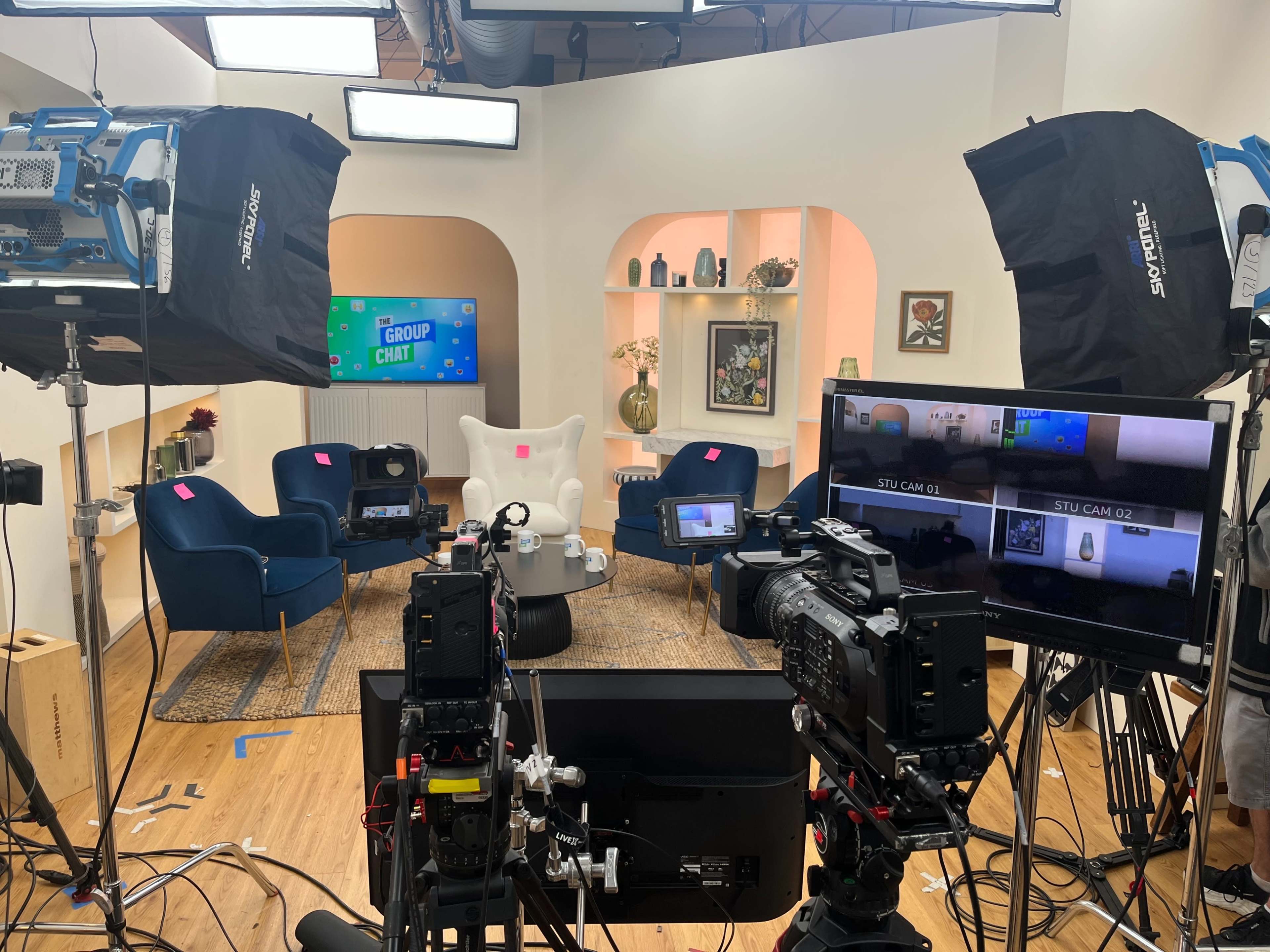 The image shows a television studio set equipped with multiple cameras, lighting equipment, and a seating arrangement.