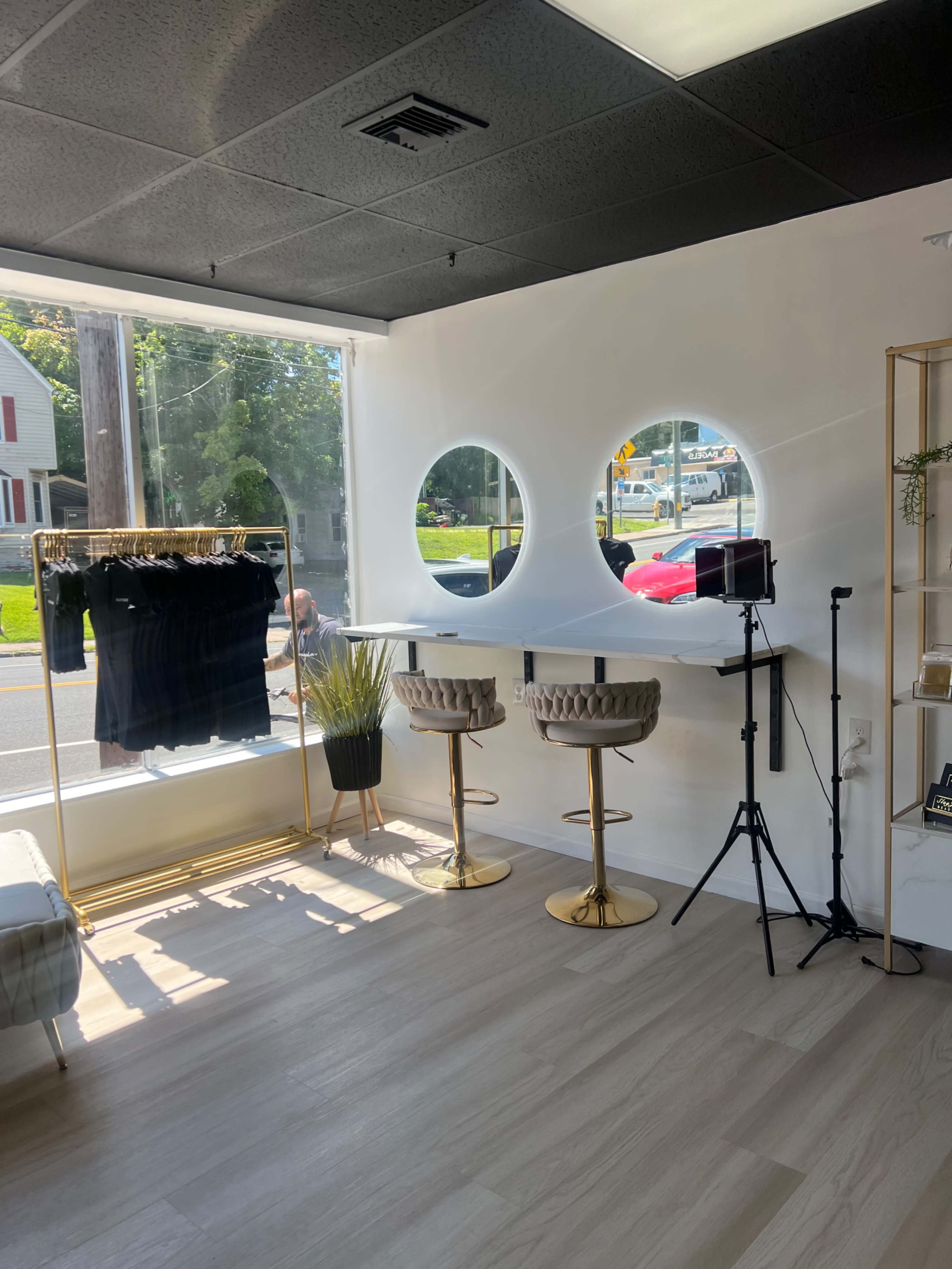 A modern retail space with two barstools, a clothing rack displaying black garments, large round windows, and a camera setup near a street view.