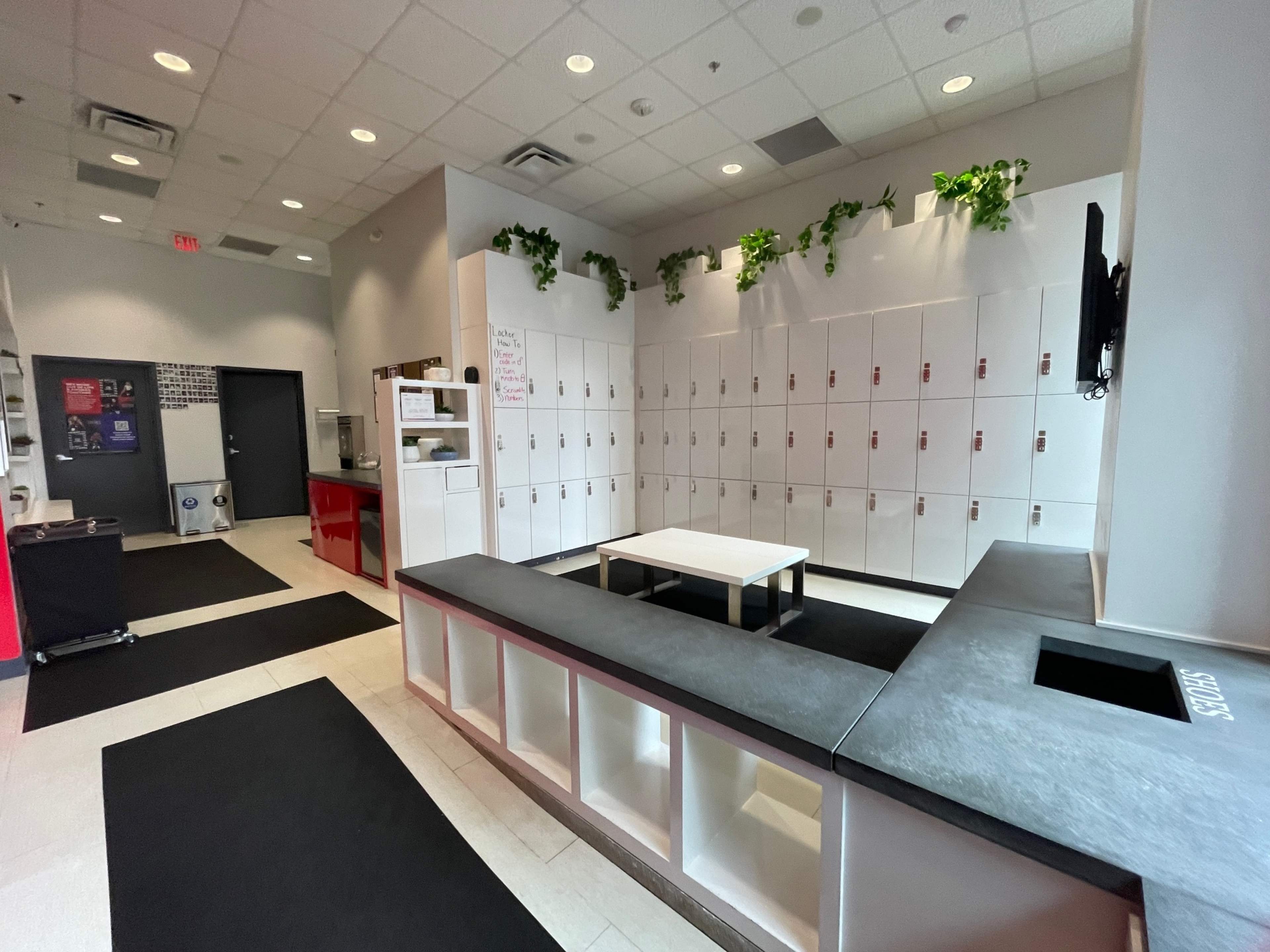 The image shows a locker room with white lockers lining the wall, a dark countertop surface, and a central seating area.