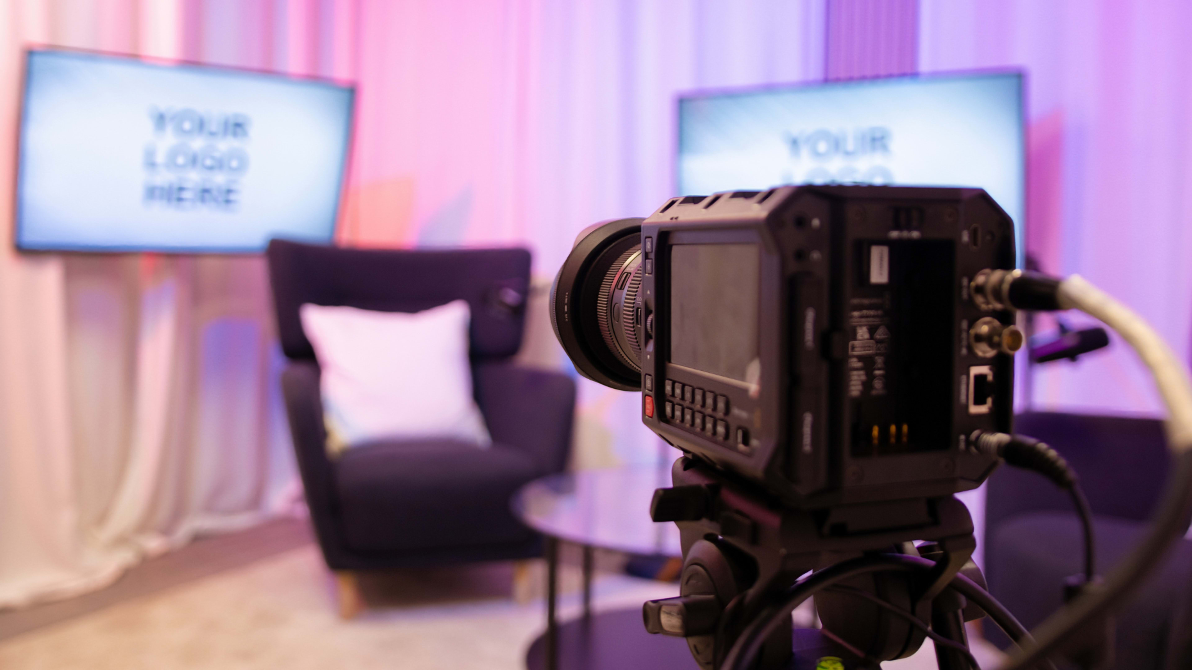 A camera is positioned in front of a cozy set featuring two screens with the text "YOUR LOGO HERE" and a stylish chair.