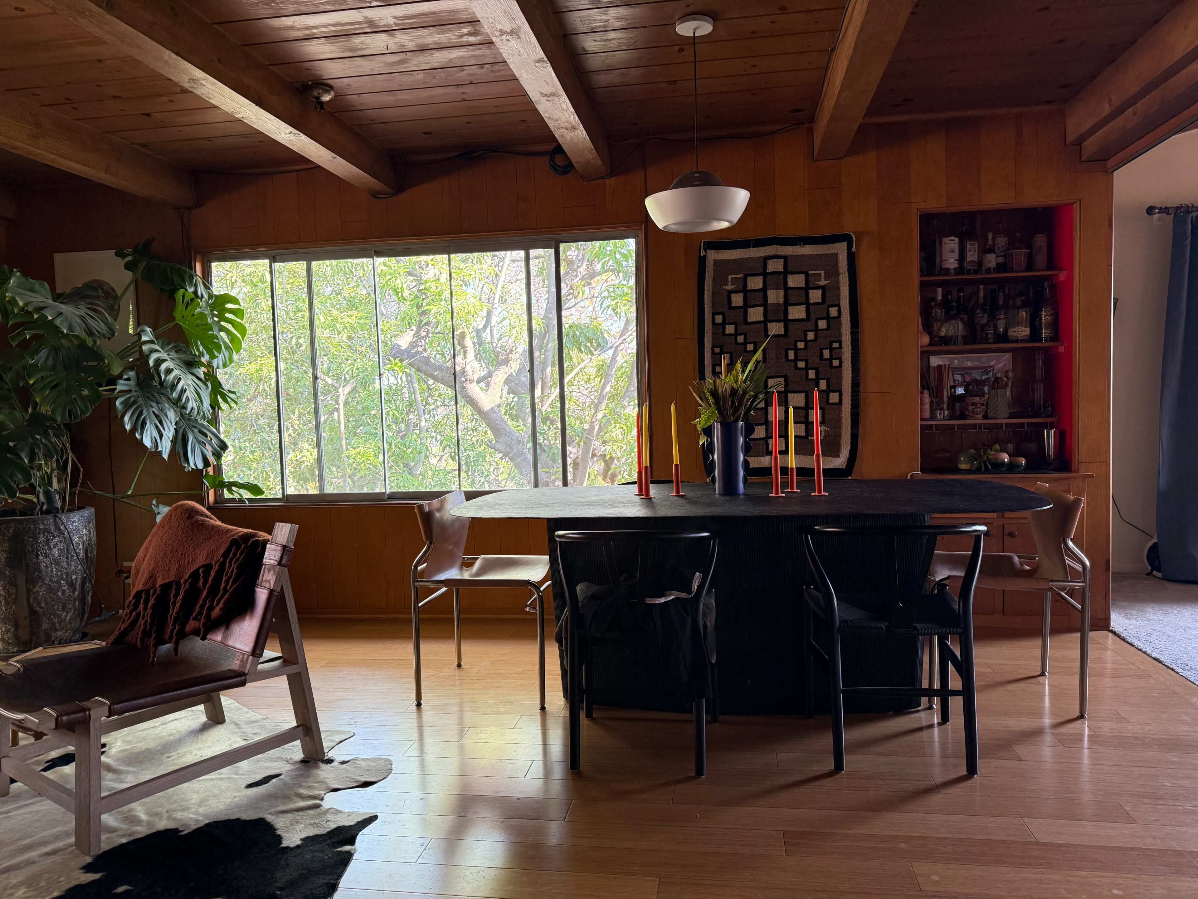 A dining area features a long black table surrounded by chairs, with a large window displaying green foliage outside.