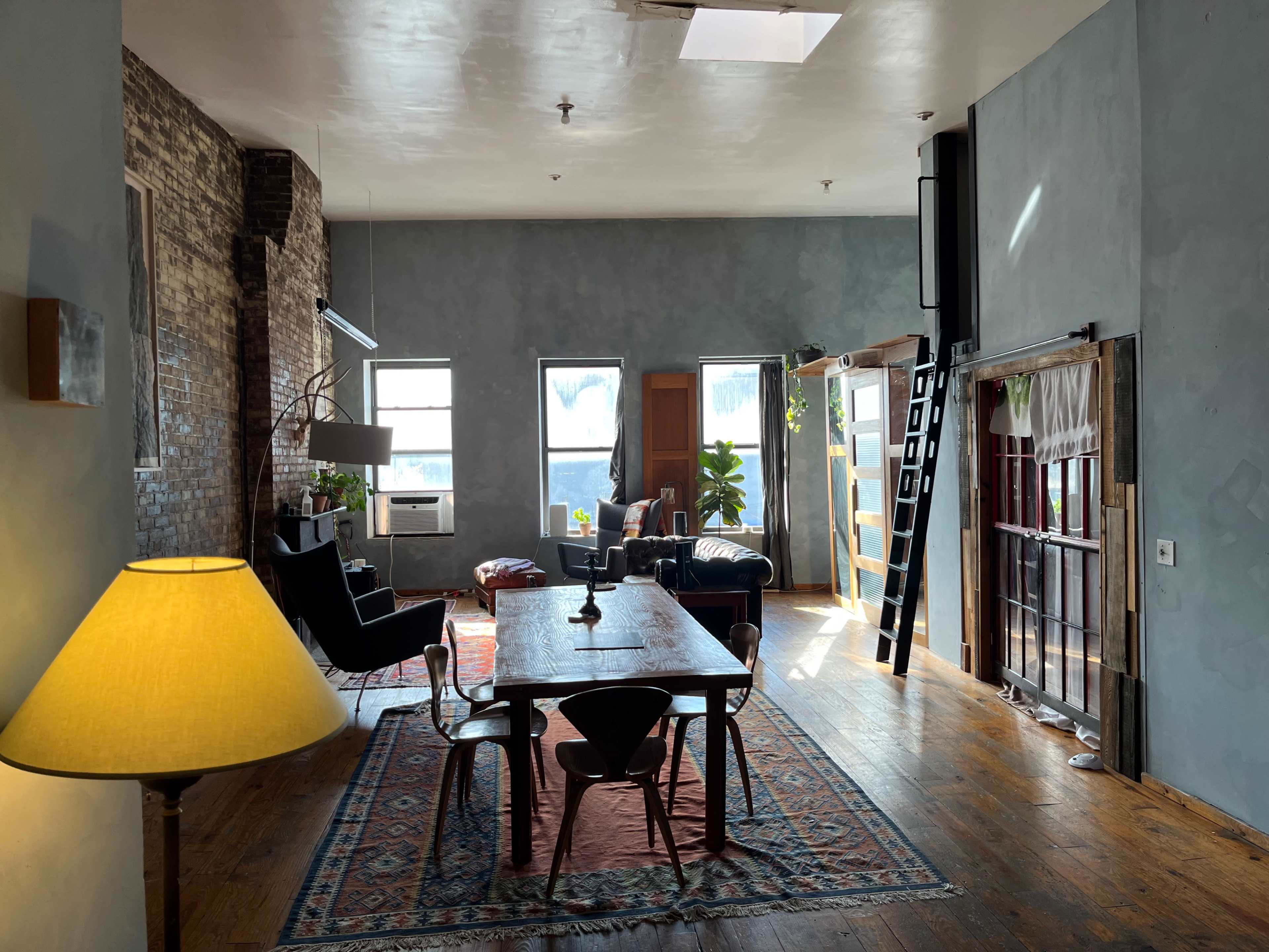 A spacious, industrial-style loft featuring a large dining table, comfortable seating arrangements, and large windows allowing natural light to fill the room.