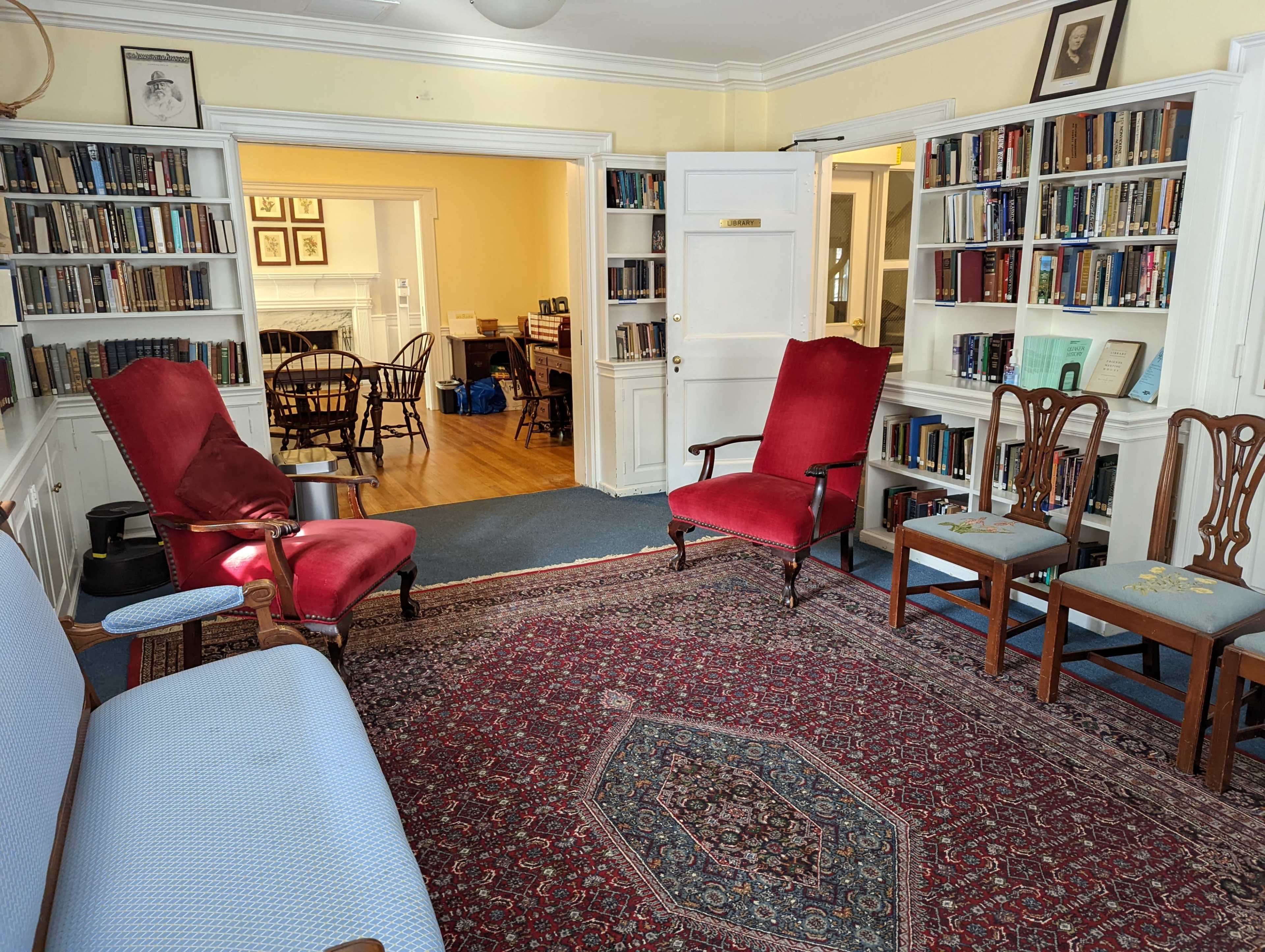A cozy library with two red upholstered chairs, a blue sofa, and several book-filled shelves lining the walls, leading into a dining area visible in the background.