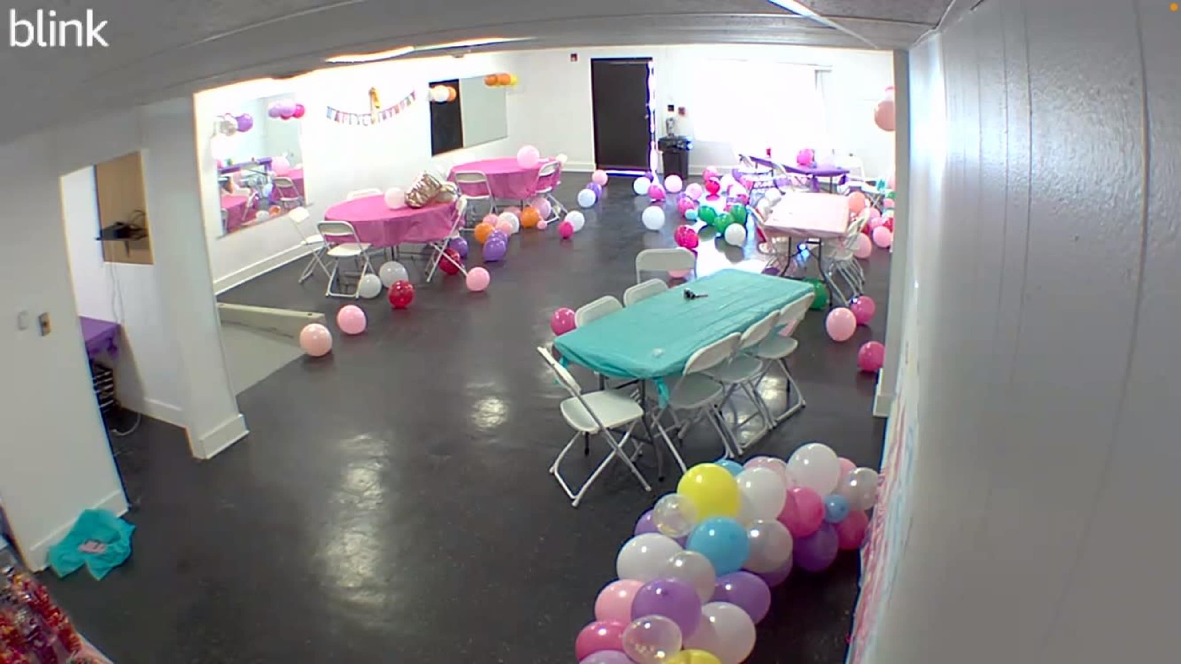 The image shows a decorated party room with tables set up, colorful balloons scattered across the floor, and a festive atmosphere.