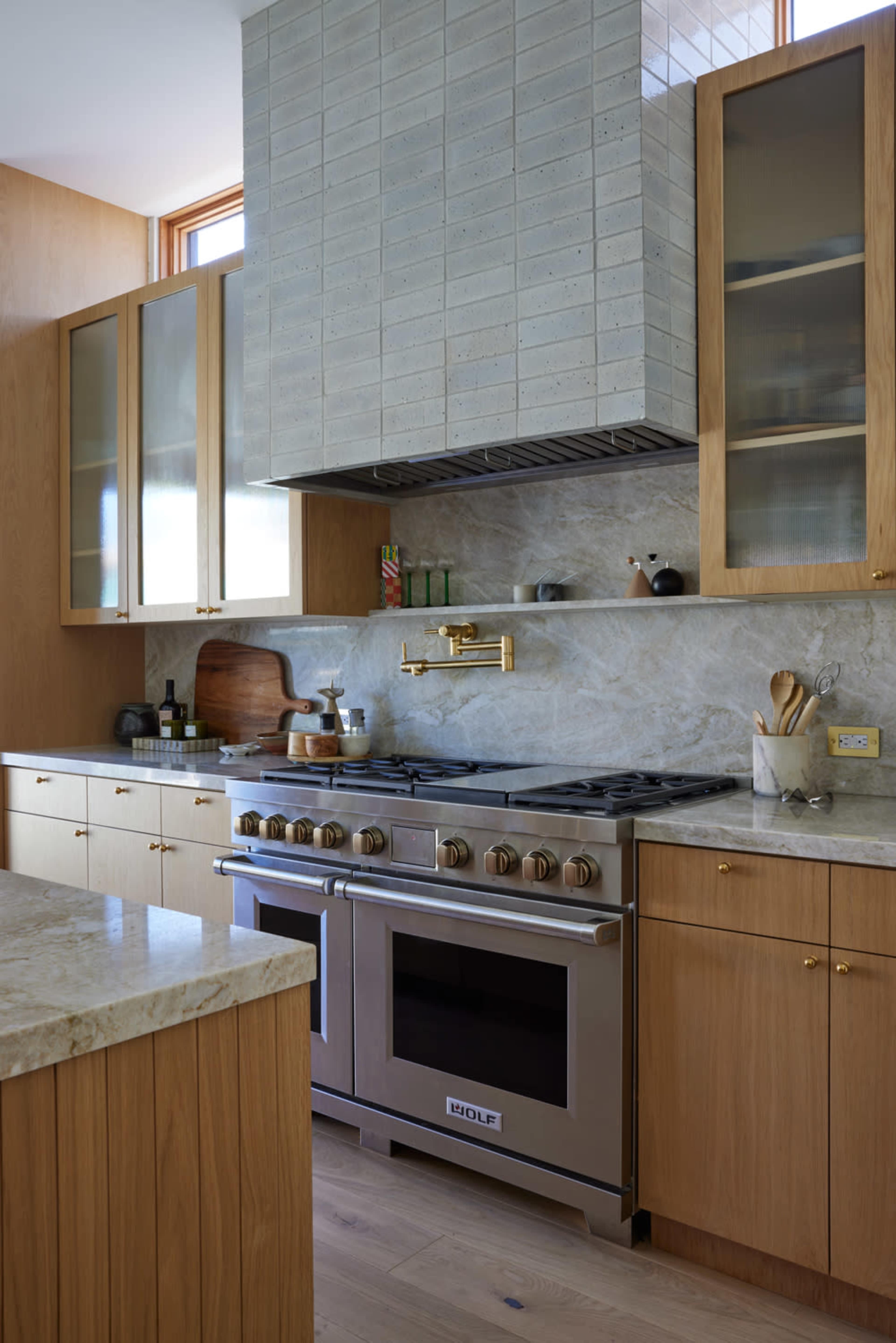 The image shows a modern kitchen with light wood cabinetry, a large stainless steel range, and a stone backsplash.