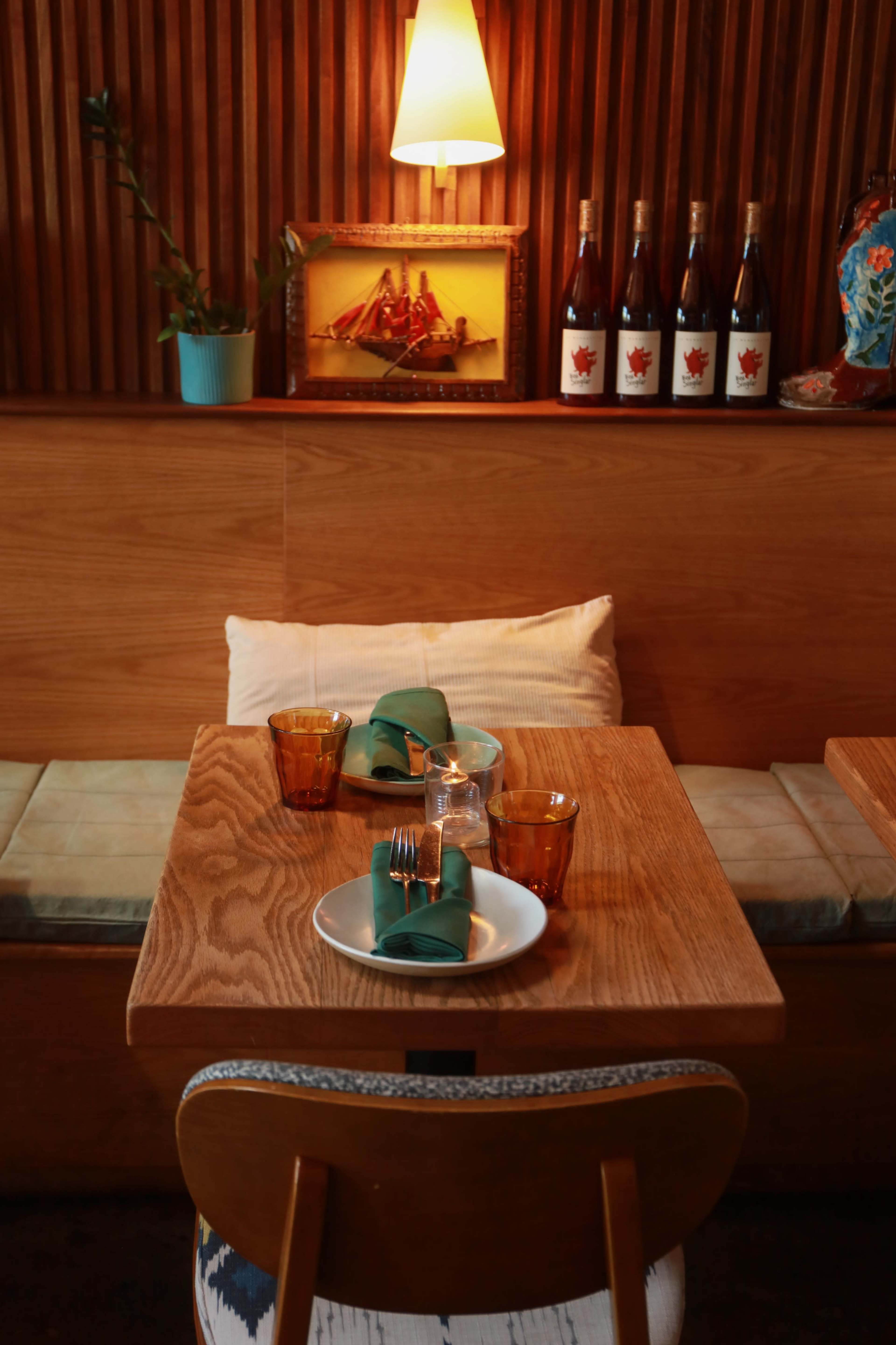 A wooden dining table is set with plates and glasses, surrounded by a cozy seating area and decorated with bottles and a framed painting on the wall.