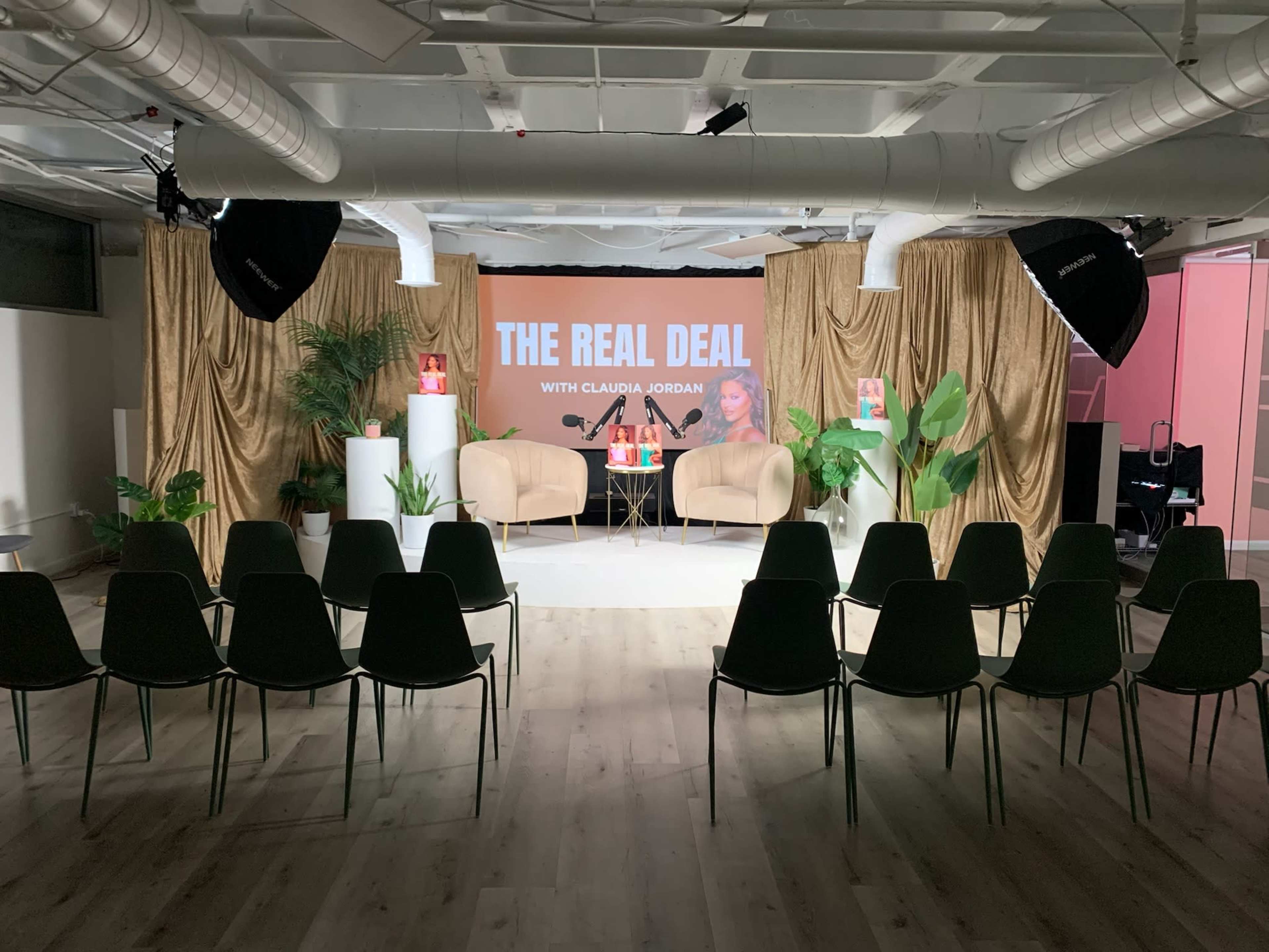 The image shows a neatly arranged event space with two chairs and a table at the front, flanked by decorative plants, and a backdrop displaying the text "THE REAL DEAL WITH CLAUDIA JORDAN."