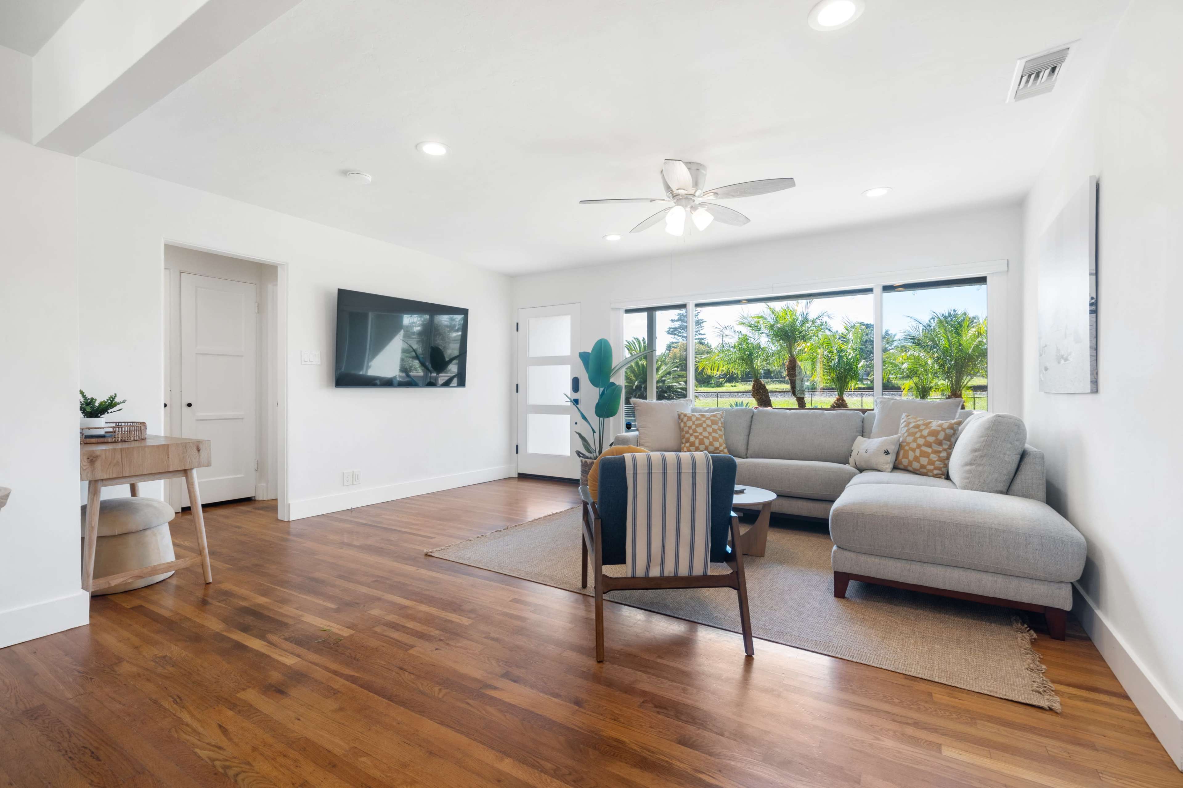 A bright living room features a gray sectional sofa, a television mounted on the wall, and large windows overlooking palm trees.