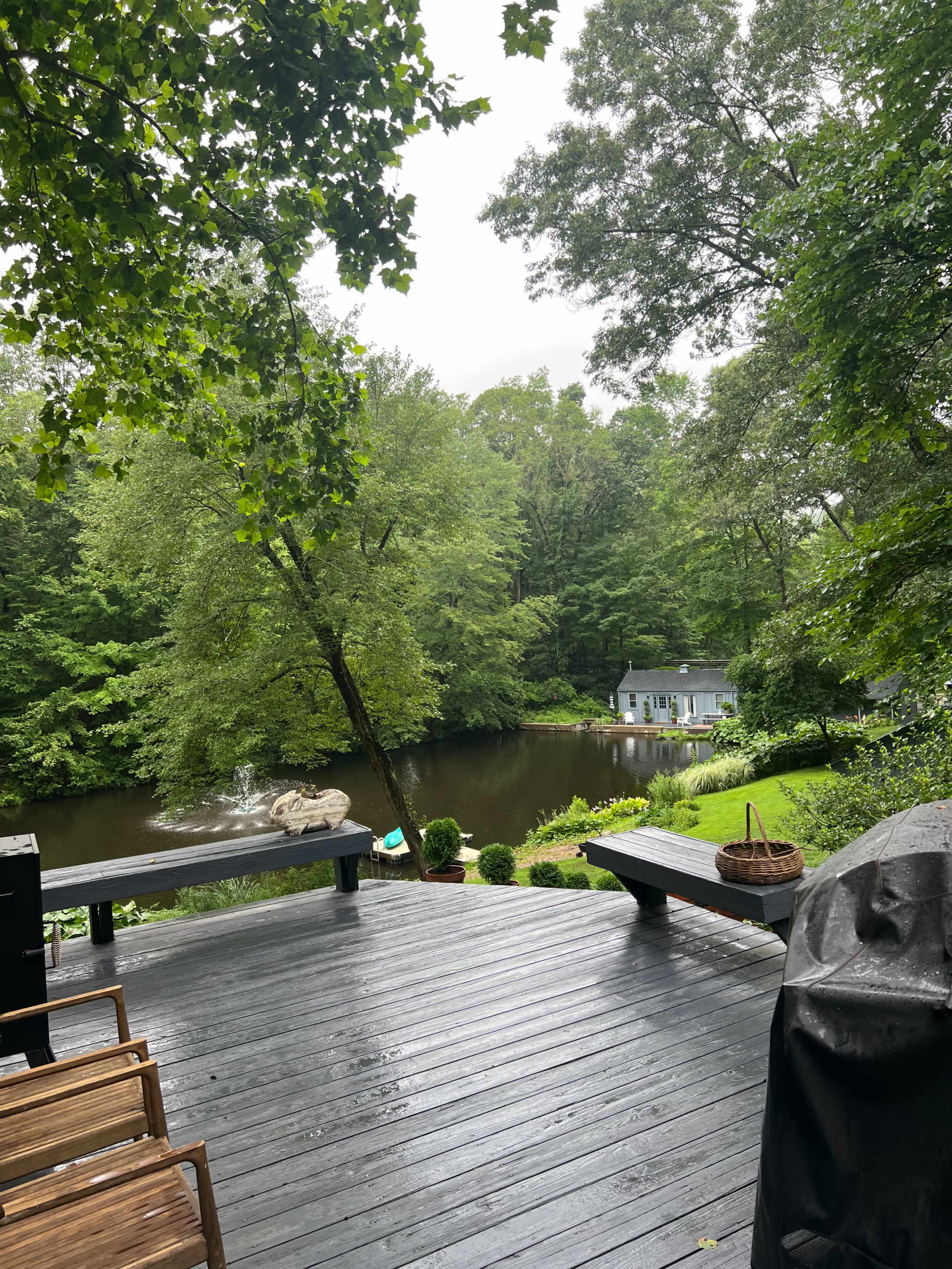 A wooden deck overlooks a tranquil pond surrounded by lush trees and a small house in the background.