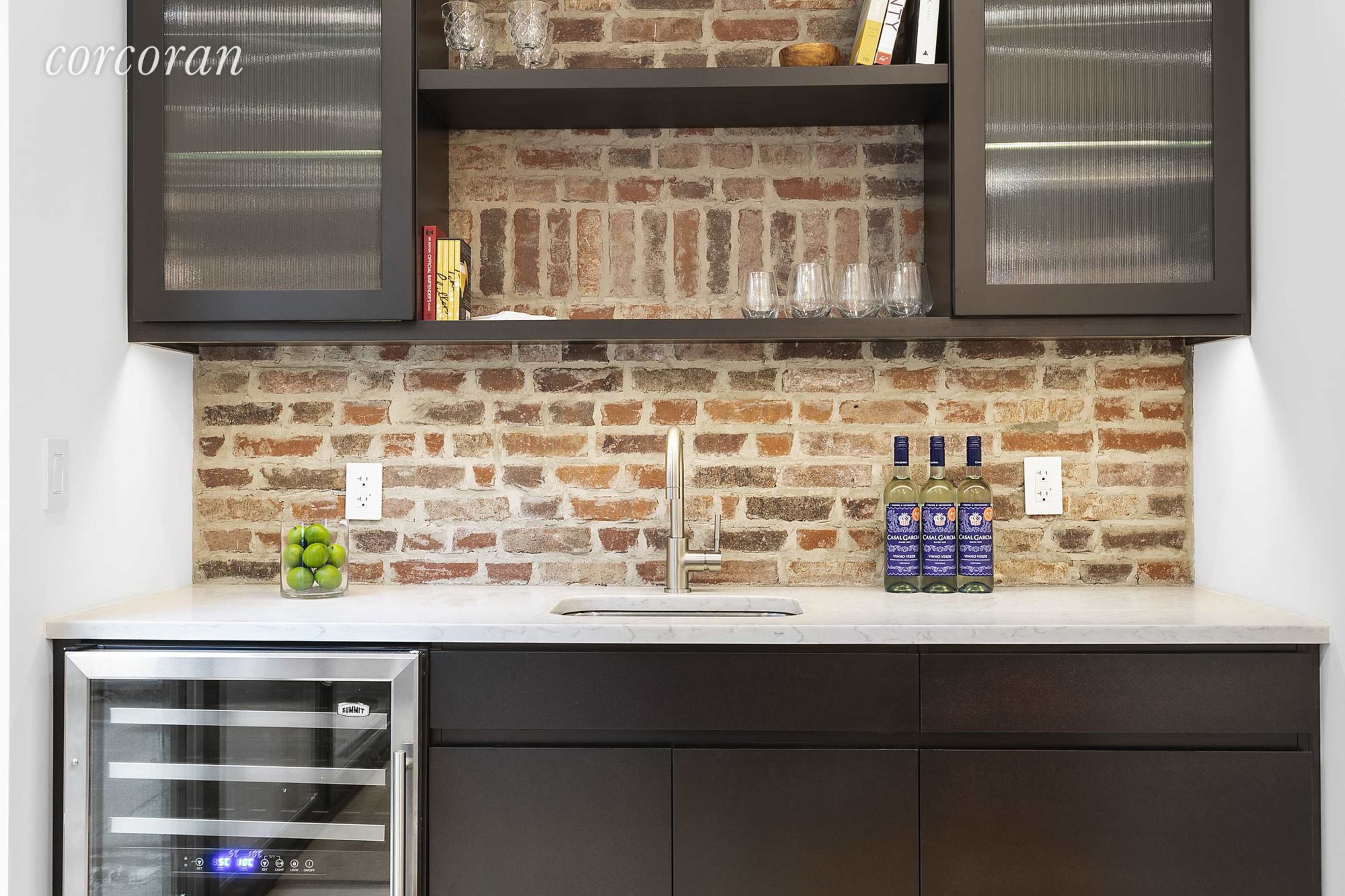 A modern kitchenette with a brick backsplash, a sink, a wine cooler, and shelves holding glasses and books.