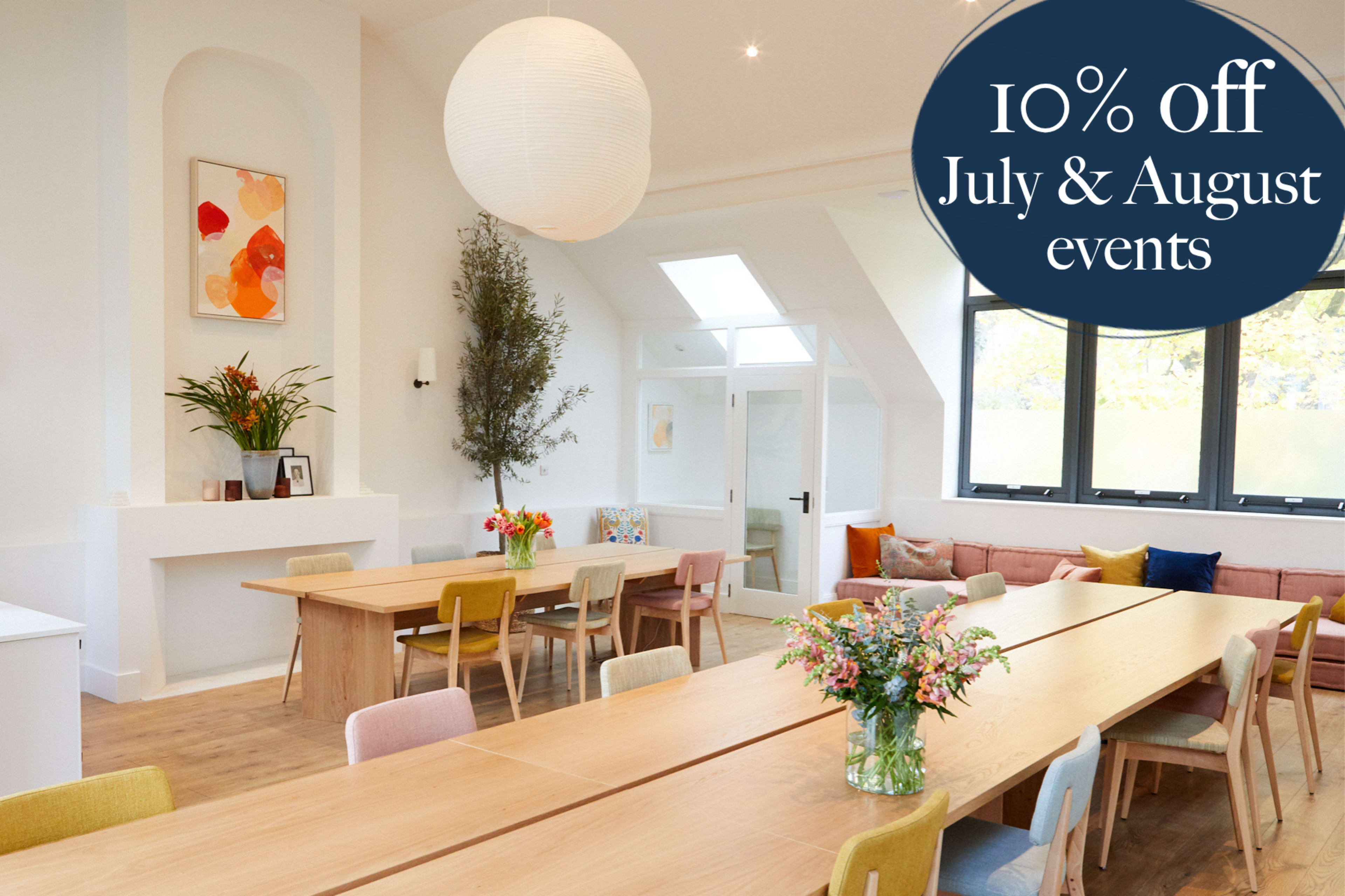 A bright and spacious event space features a long wooden table, colorful chairs, and large windows, complemented by floral arrangements and decorative artwork.