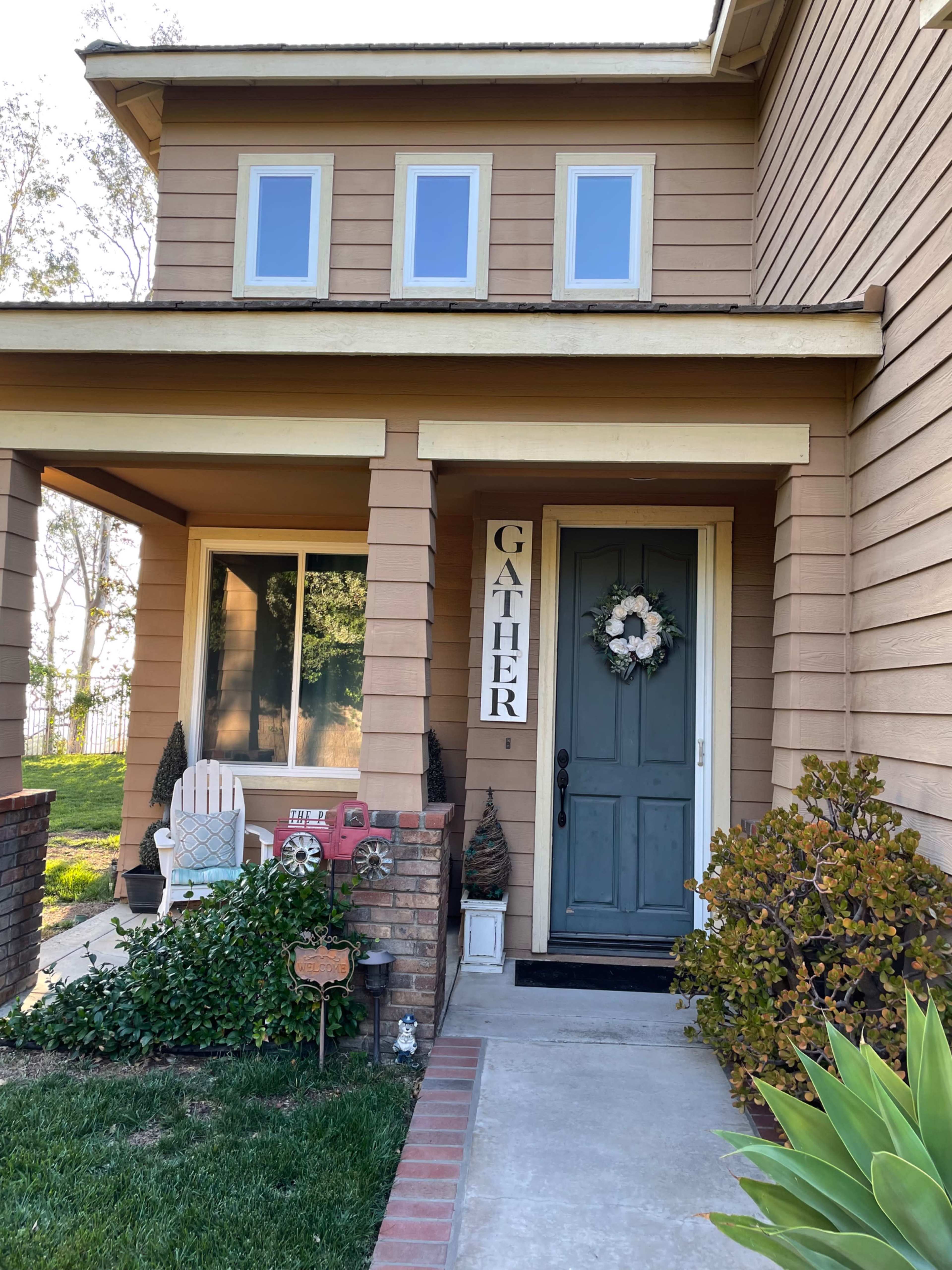 The front entrance of a house features a blue door, a large "GATHER" sign, a wreath, and a small seating area with patio furniture.