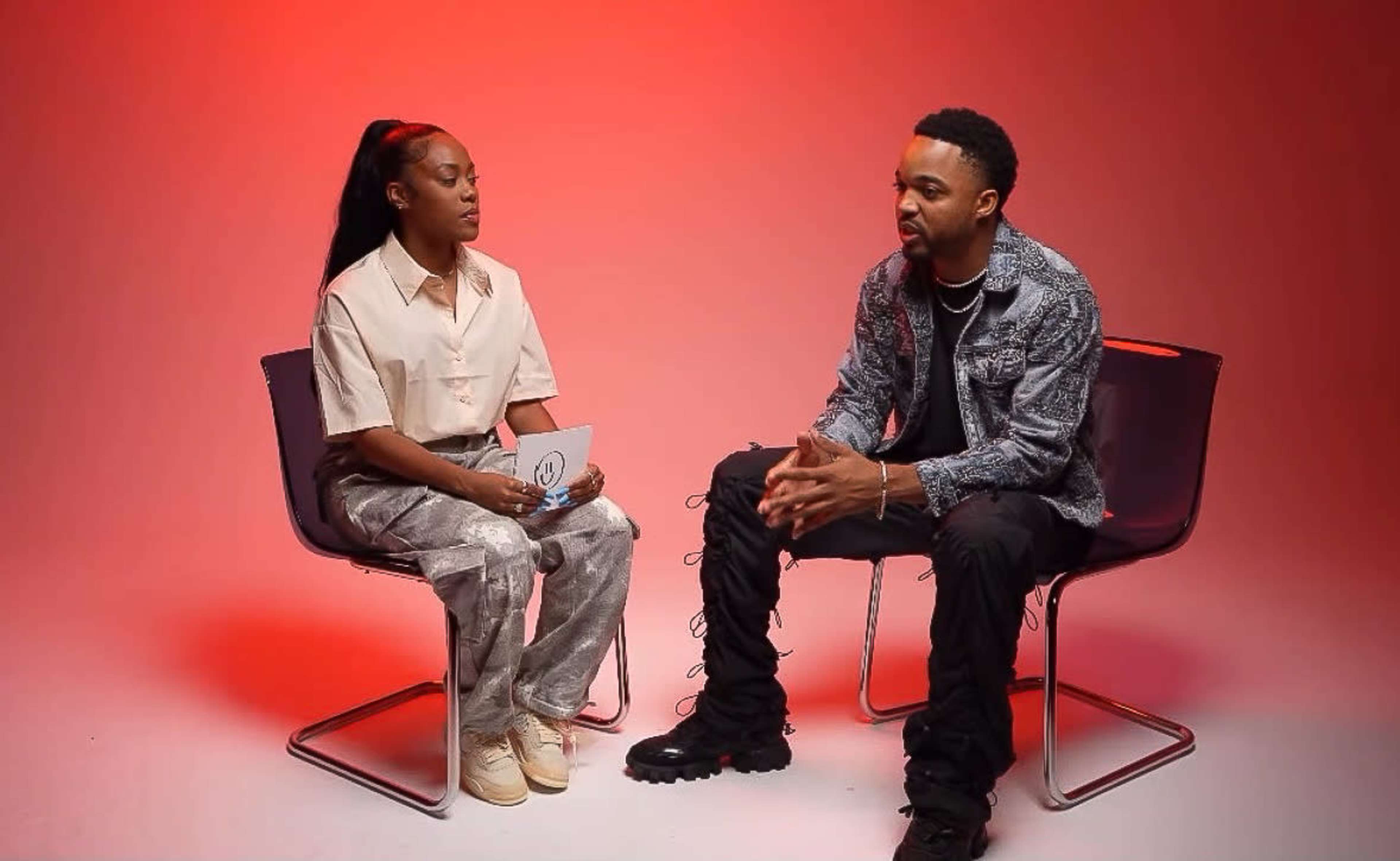 A man and a woman are seated in modern chairs, facing each other against a red backdrop during an interview.