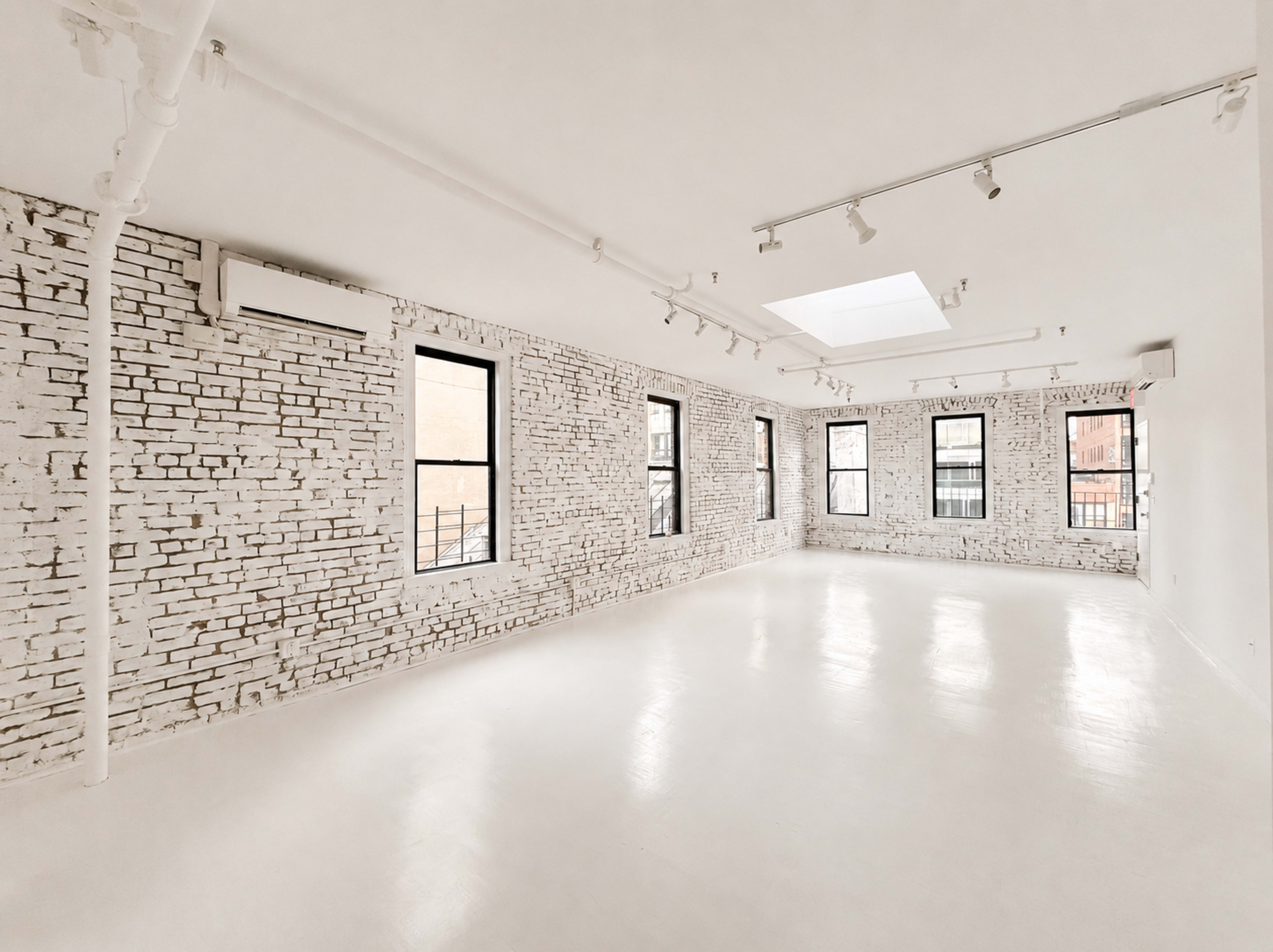 The image shows a spacious, sunlit room with whitewashed brick walls and large windows, featuring a smooth white floor and overhead lighting.