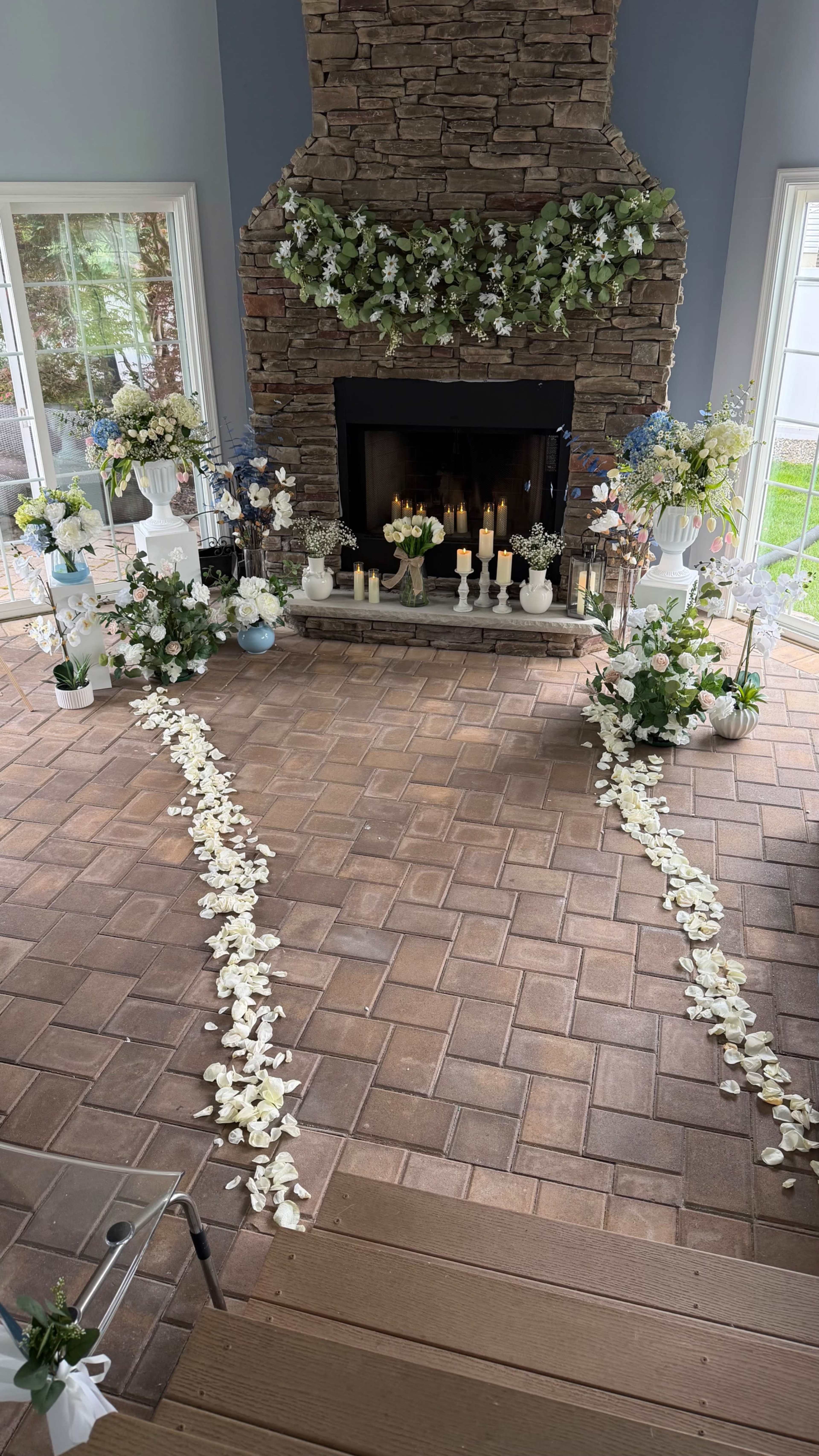 The image shows a decorated indoor space with a stone fireplace, surrounded by floral arrangements and a pathway lined with white petals leading to the fireplace.