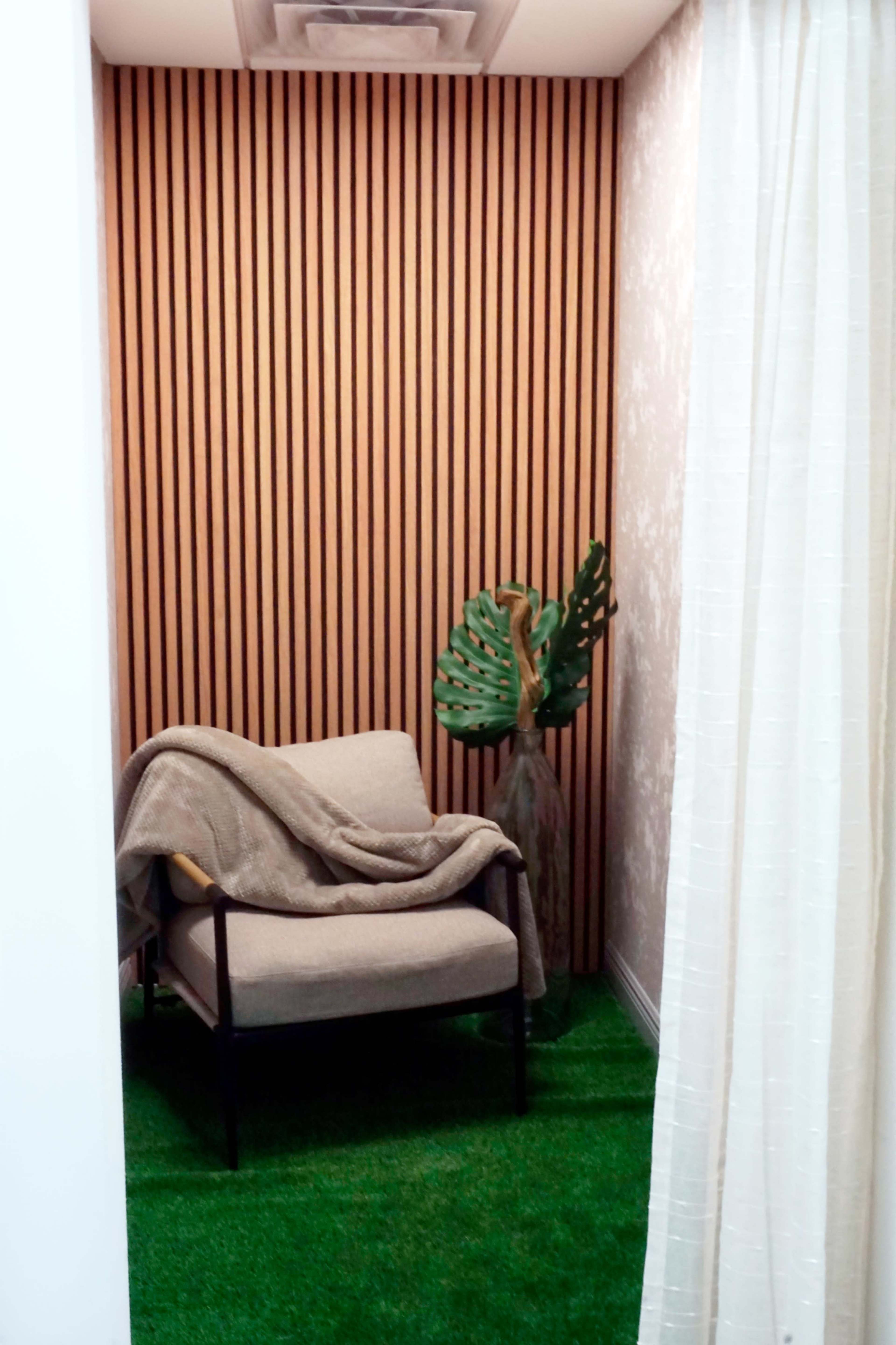 The image shows a small nook featuring a chair draped with a blanket, a decorative plant, and a backdrop of wooden paneling and artificial grass.