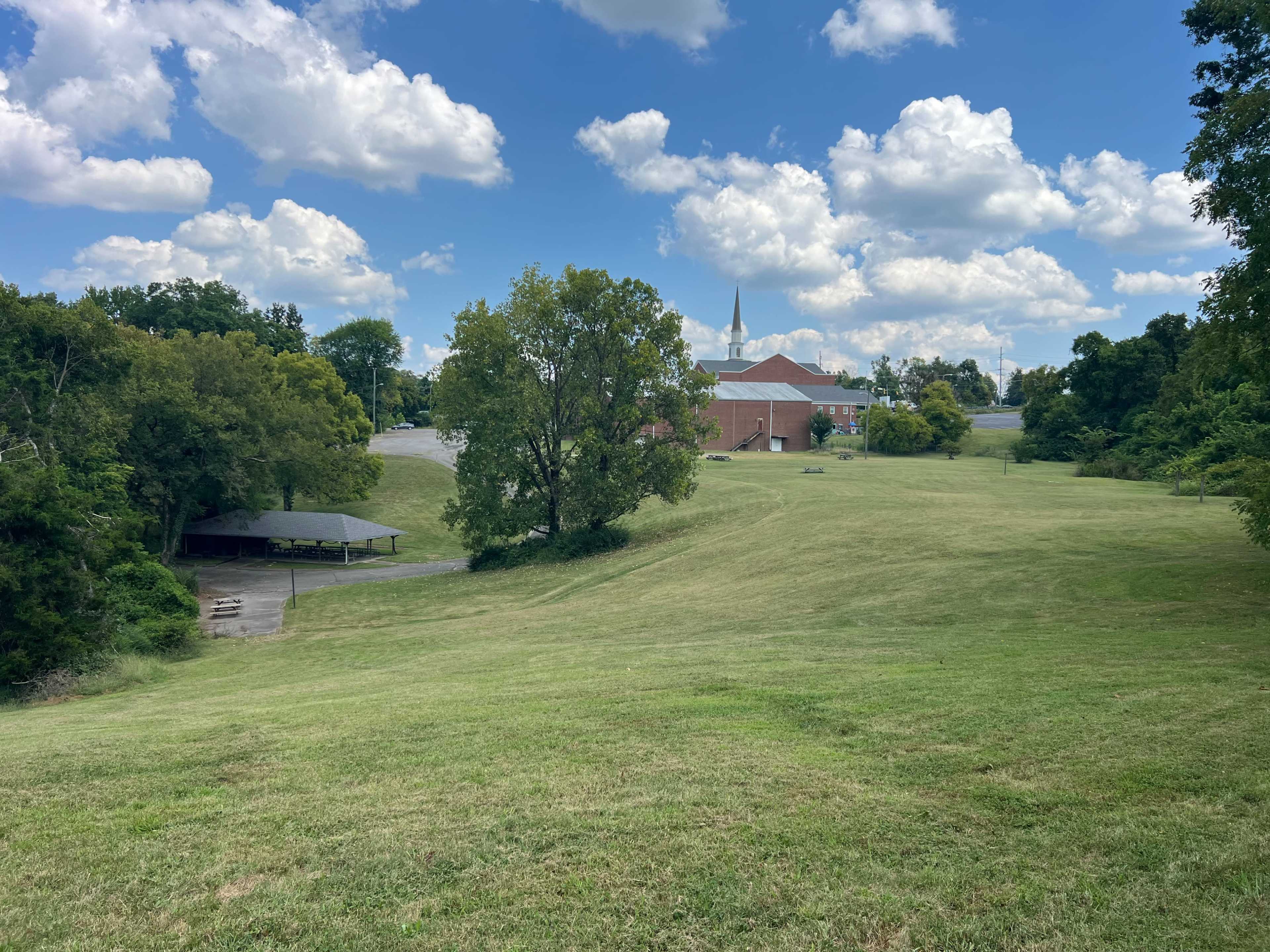 The image shows a wide green field with a few trees in the foreground and a building, possibly a church, in the background under a partly cloudy sky.
