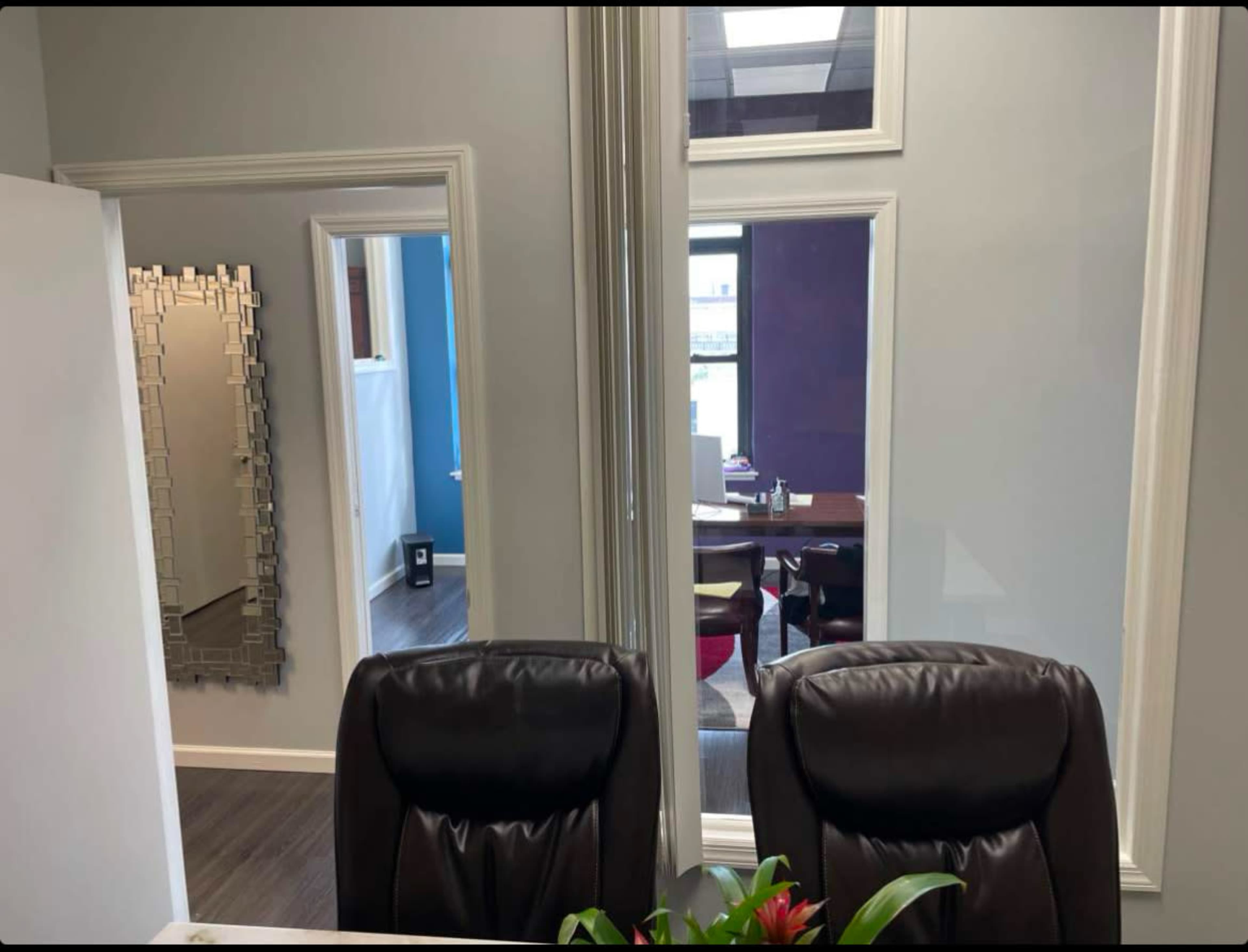 The image shows an office space with two leather chairs facing a table, a large mirror on the left wall, and a view of a brightly colored room through a window.