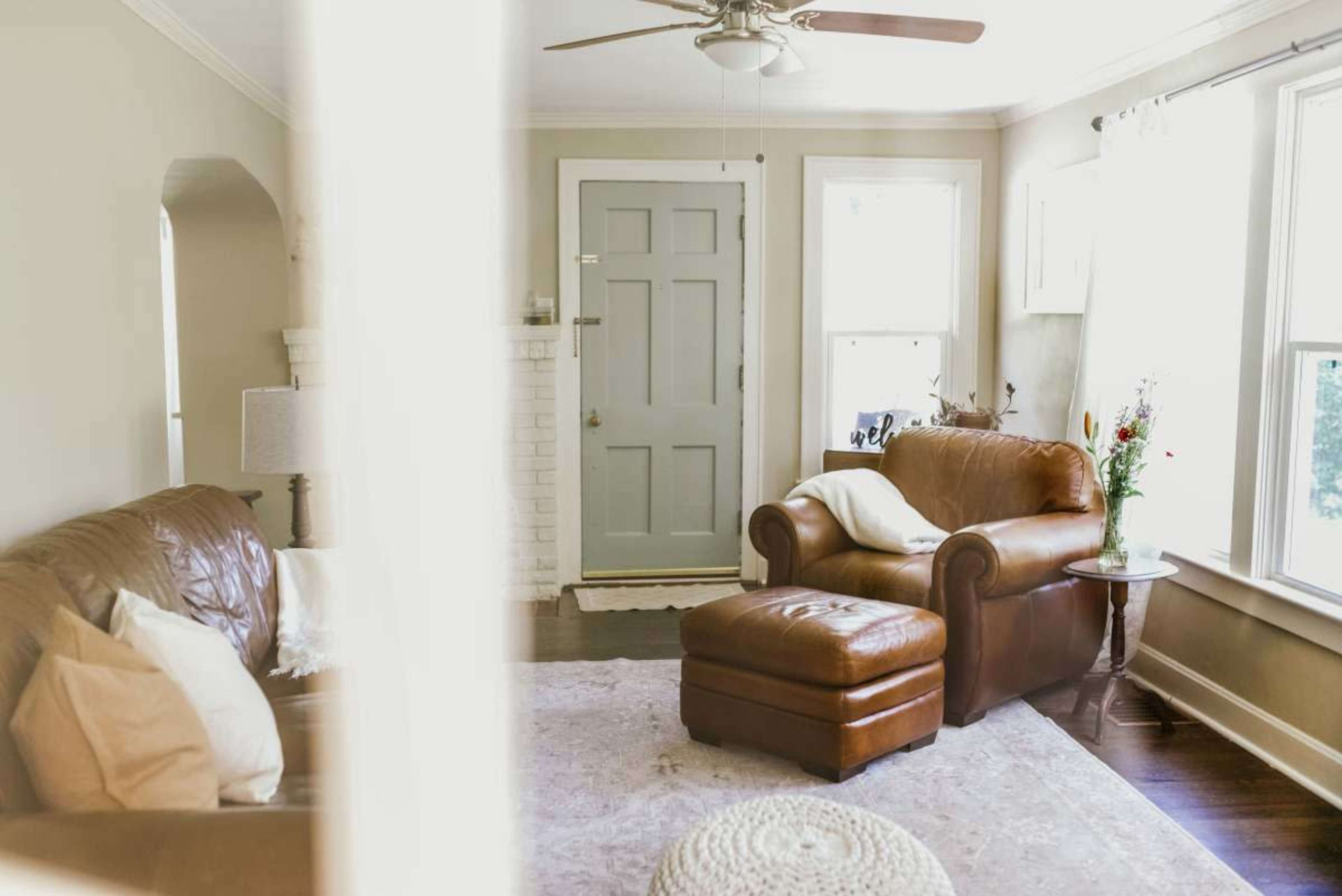 A cozy living room features a brown leather sofa and armchair, with a small side table and natural light streaming through the windows.