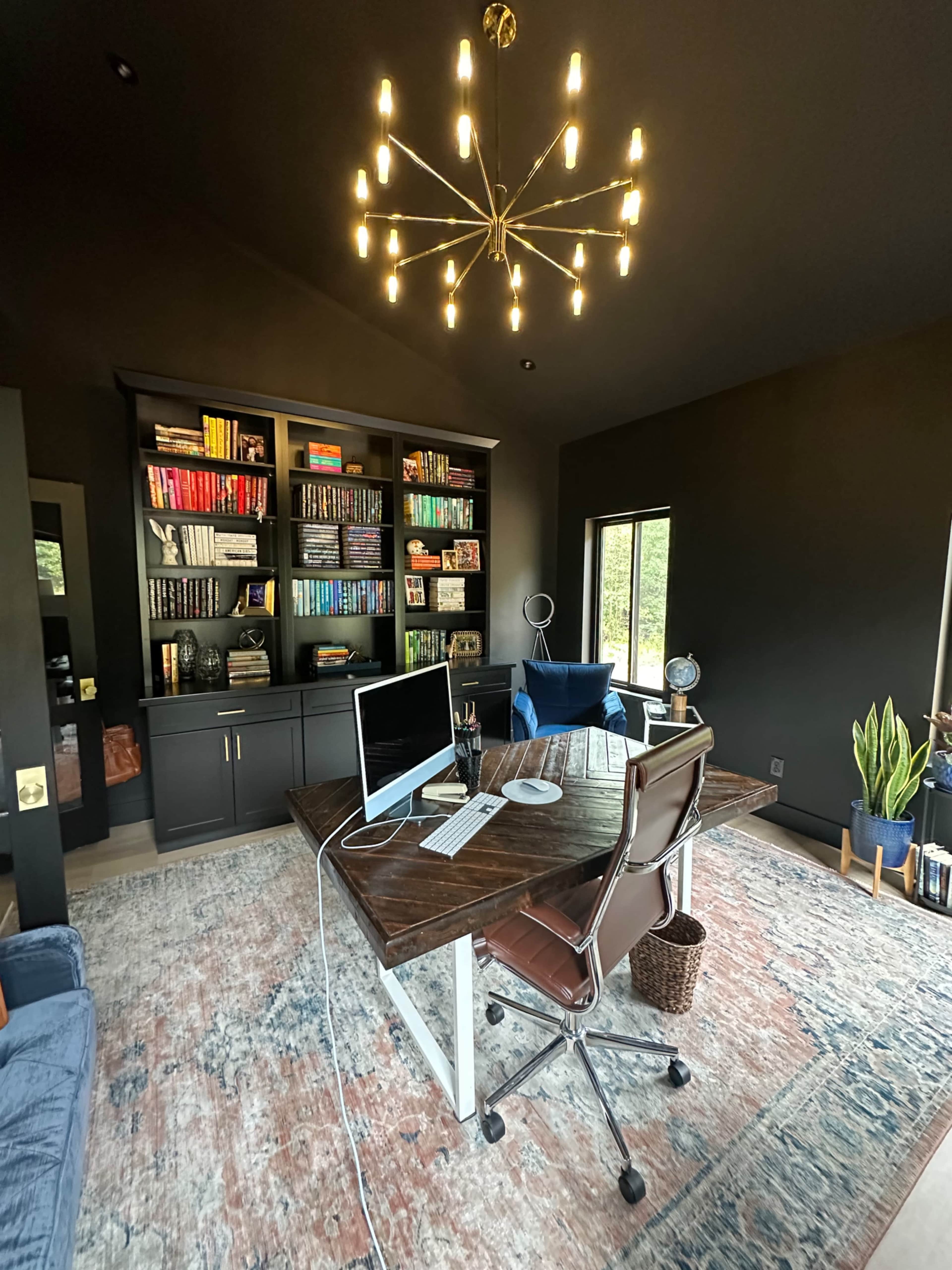 The image shows a modern office space featuring a wooden desk, an ergonomic chair, a large bookshelf filled with books, and a decorative chandelier overhead.