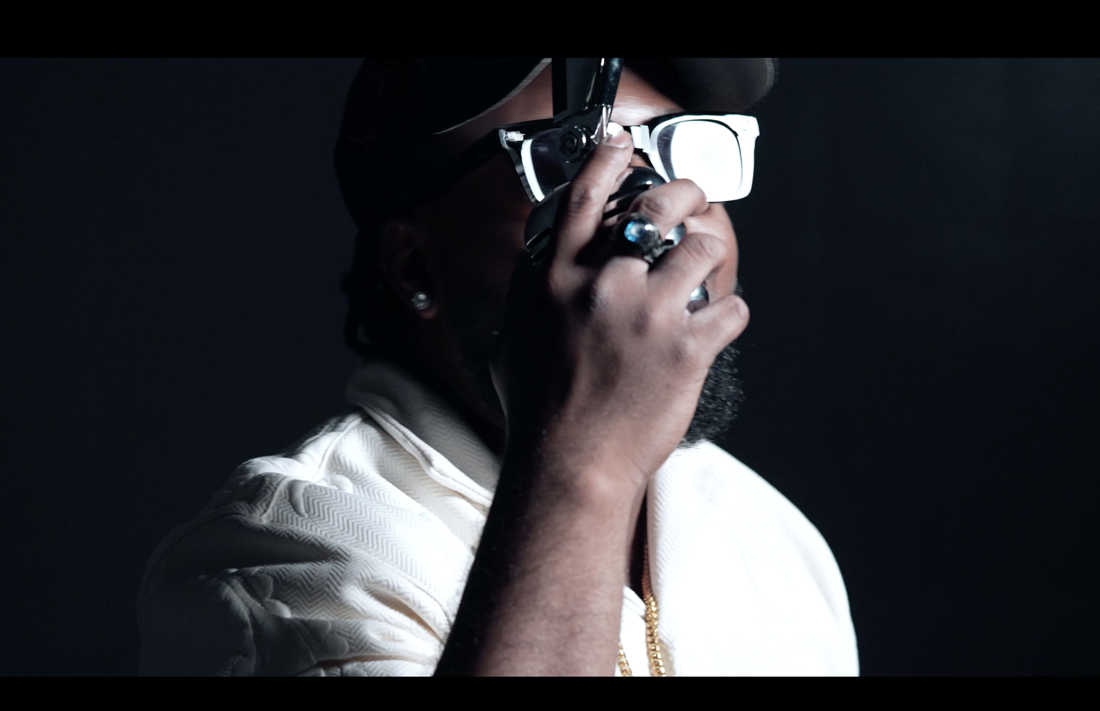 A person wearing sunglasses and holding a camera poses in a dimly lit setting.