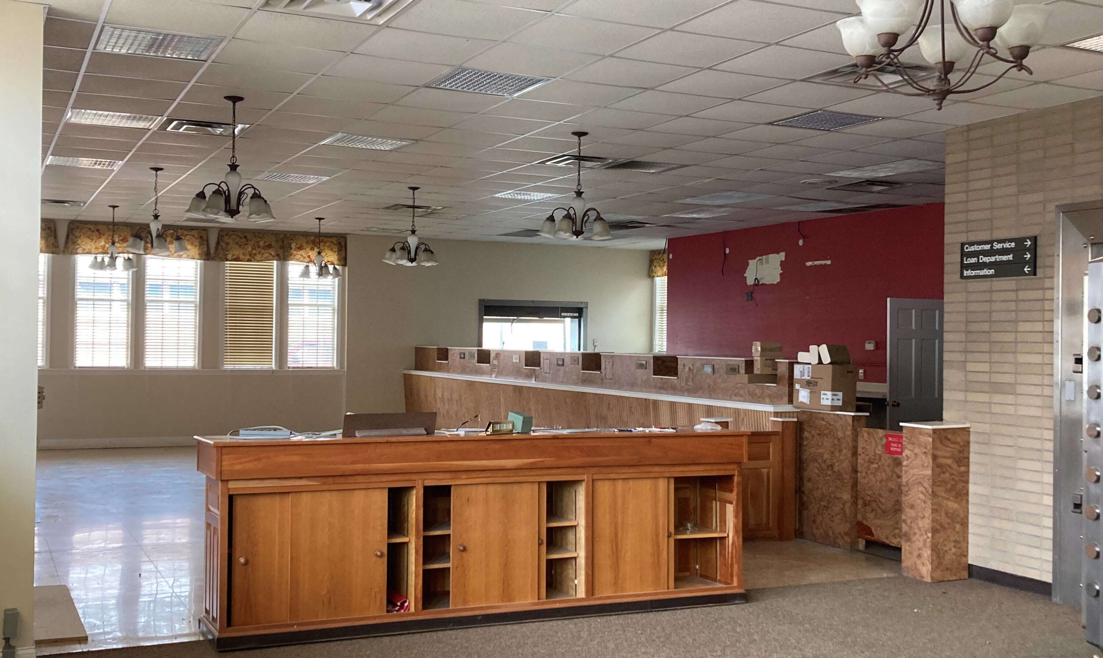 An empty reception area with a wooden counter and rows of cubicles, featuring large windows and a red accent wall.