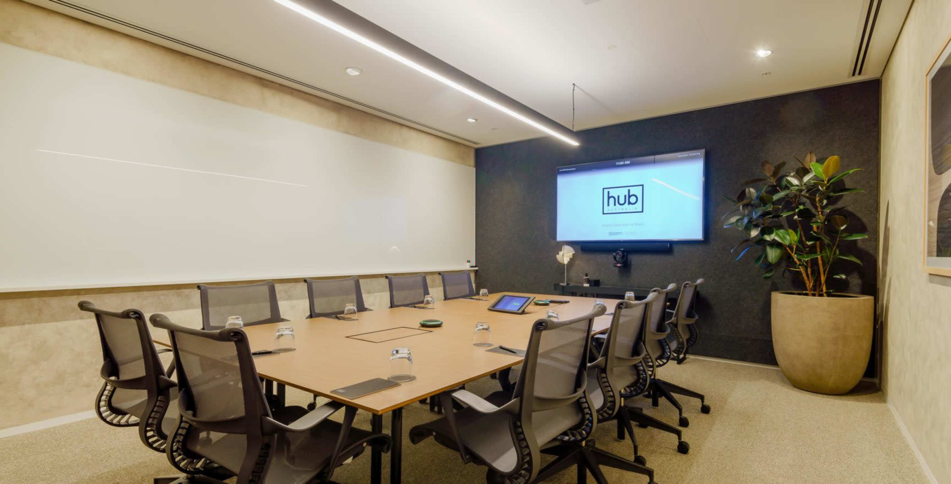 A modern conference room featuring a large table surrounded by office chairs, a wall-mounted screen, and a plant in a pot.