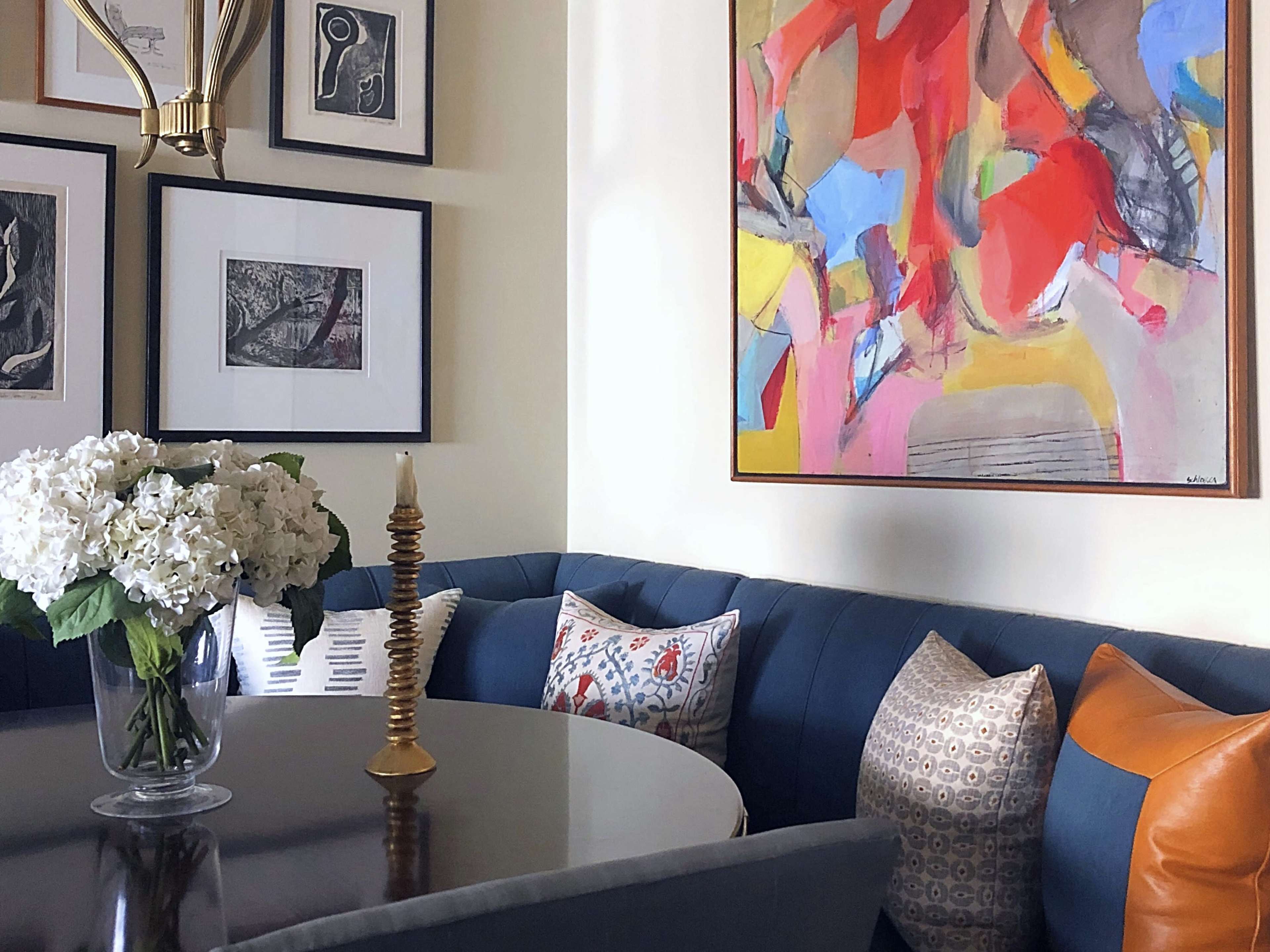 A corner of a room features a round dark table surrounded by a blue upholstered banquette, adorned with a vase of hydrangeas and several decorative pillows, alongside framed artwork on the walls.