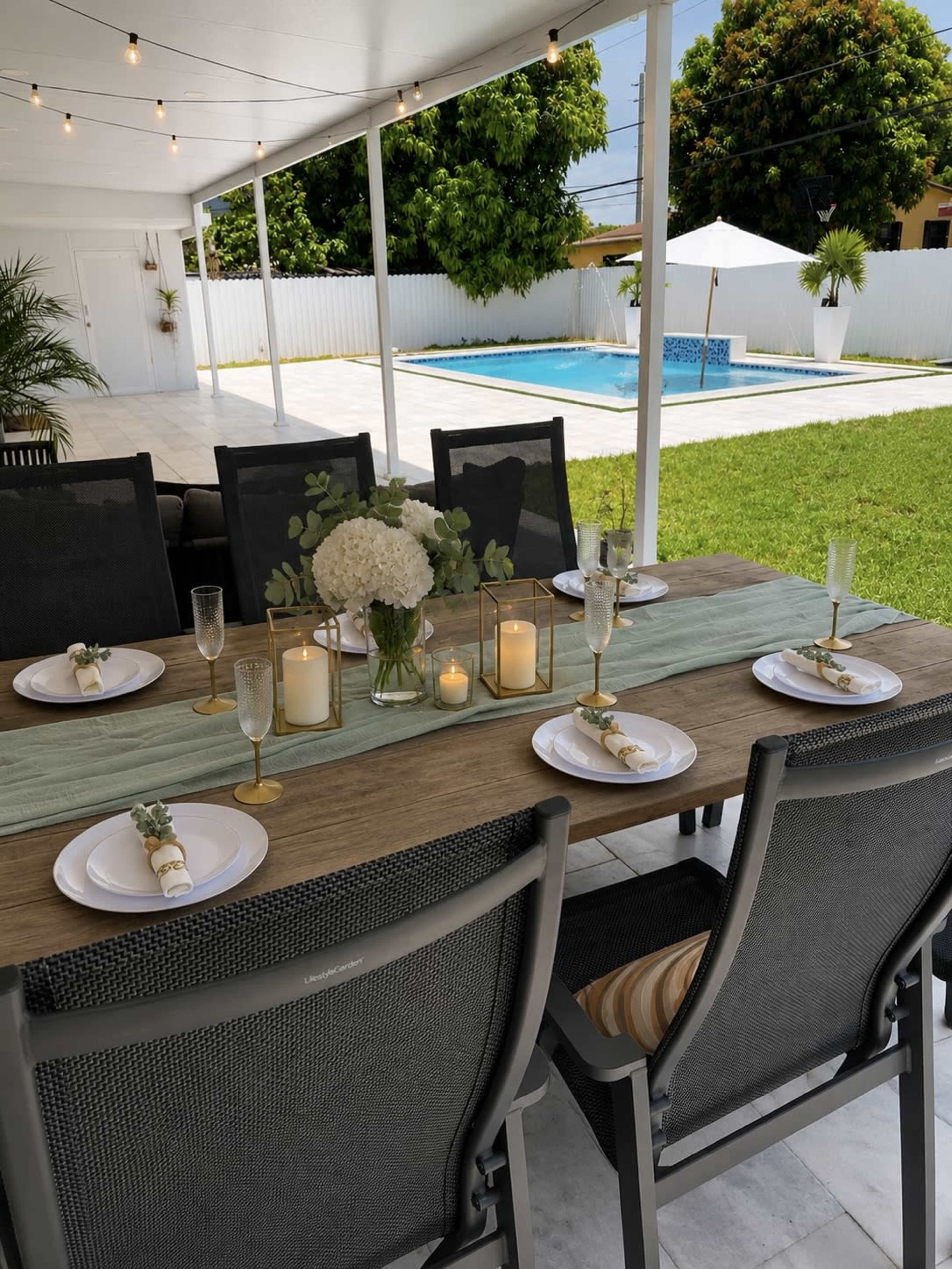 A dining table is set with plates and candles under a covered patio, overlooking a grassy yard and a swimming pool in the background.