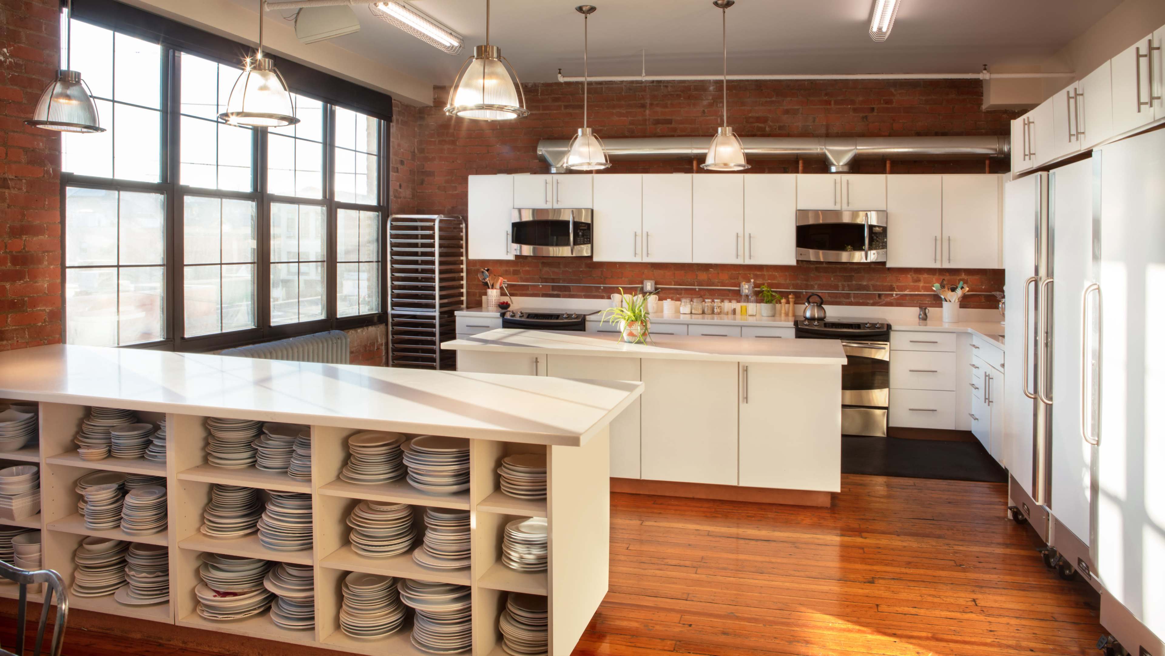 A modern kitchen with a long white island featuring open shelving filled with plates, stainless steel appliances, and large windows revealing a brick wall.