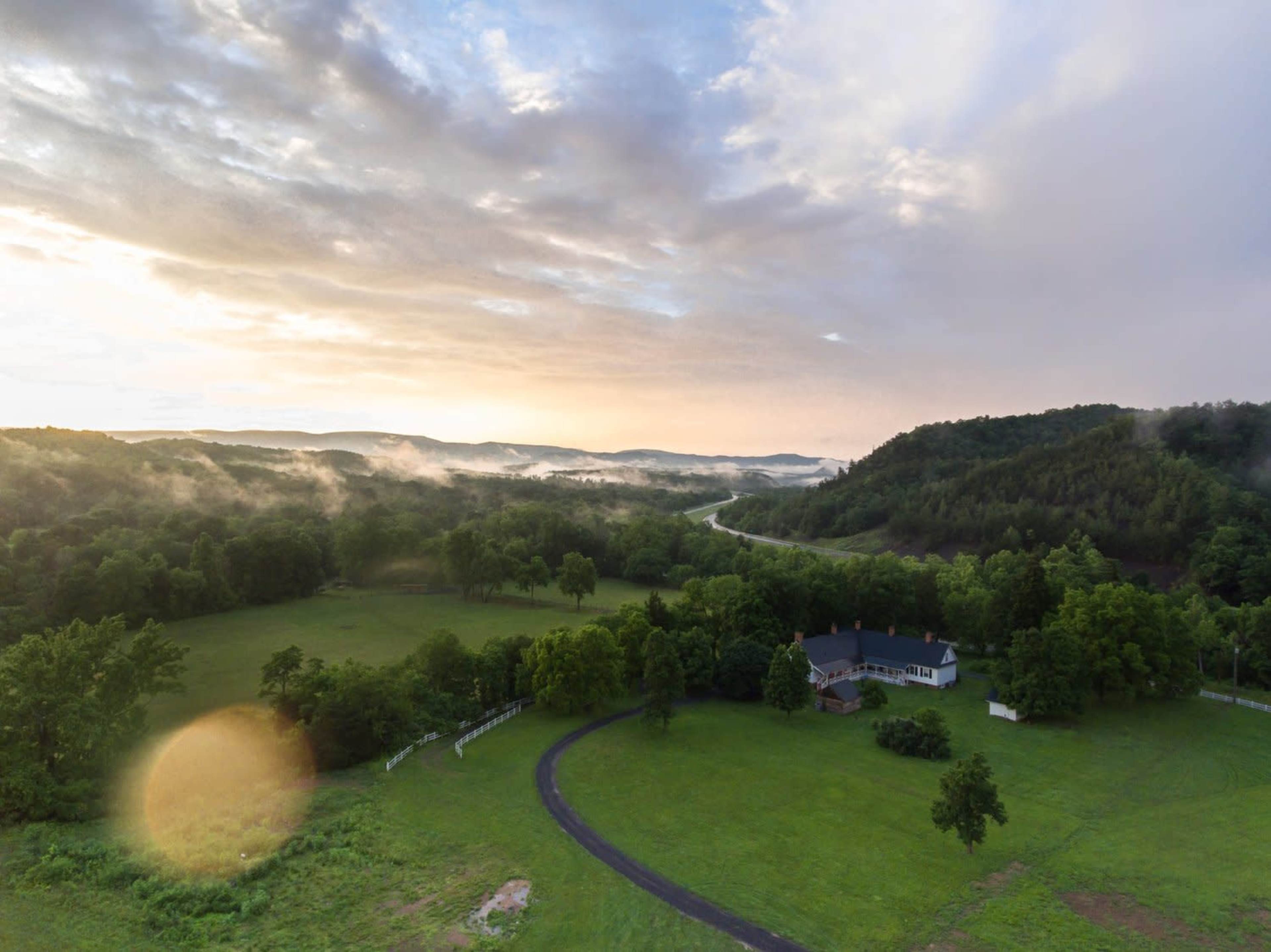 A house is situated near a winding road, surrounded by lush greenery and misty hills under a cloudy sky at dawn.